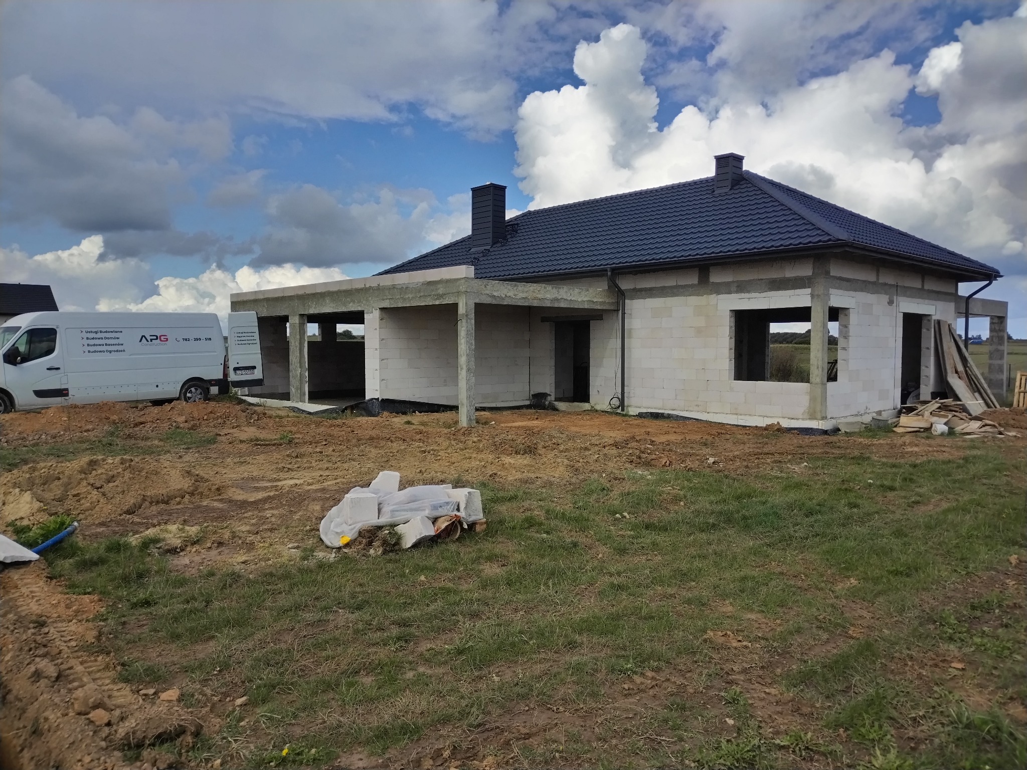 Budowa domu z białego bloczka, widoczna część dachu z czarną dachówką. Na posesji stoi firmowy bus APG Construction. Niebo z chmurami.