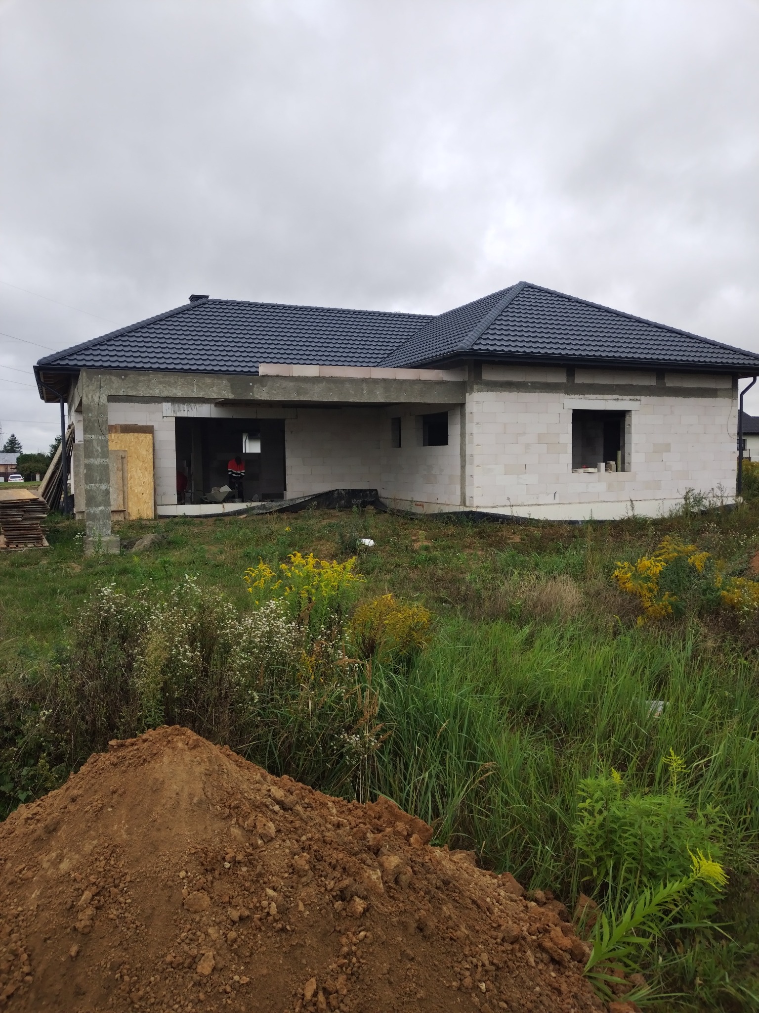 Niedokończony dom z białych bloczków, z ciemnym dachem, na działce z bujną roślinnością i stertą ziemi na pierwszym planie. Wnętrze w stanie surowym.