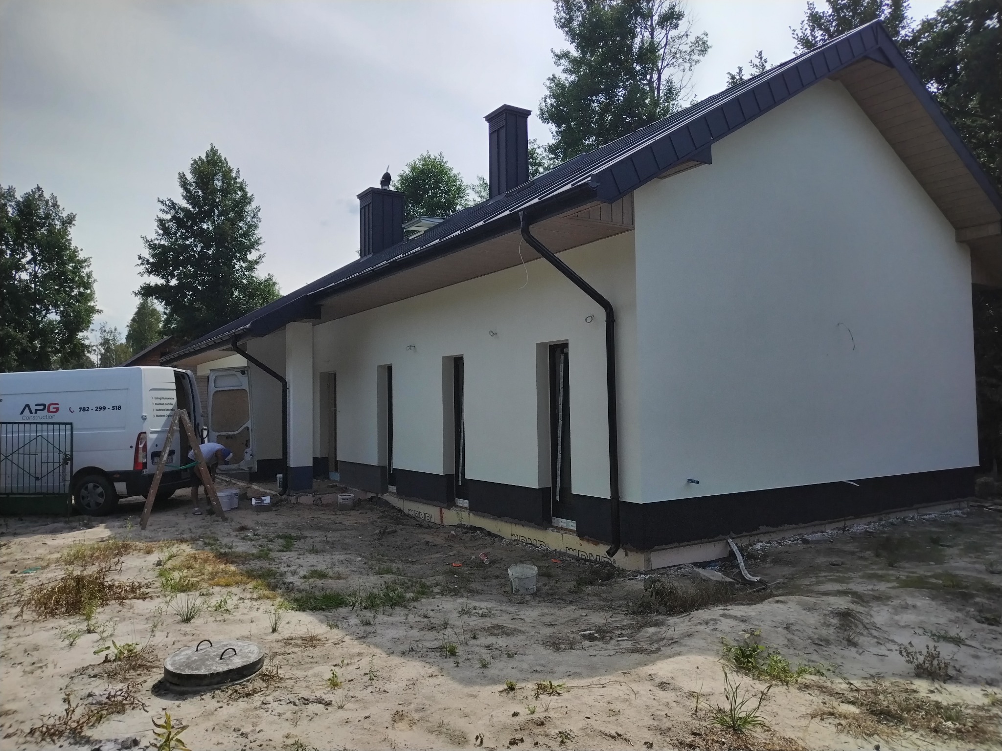 Elewacja domu jednorodzinnego w trakcie prac wykończeniowych. Widoczny fragment ocieplenia fundamentów, biała elewacja i pracownik przy samochodzie dostawczym.