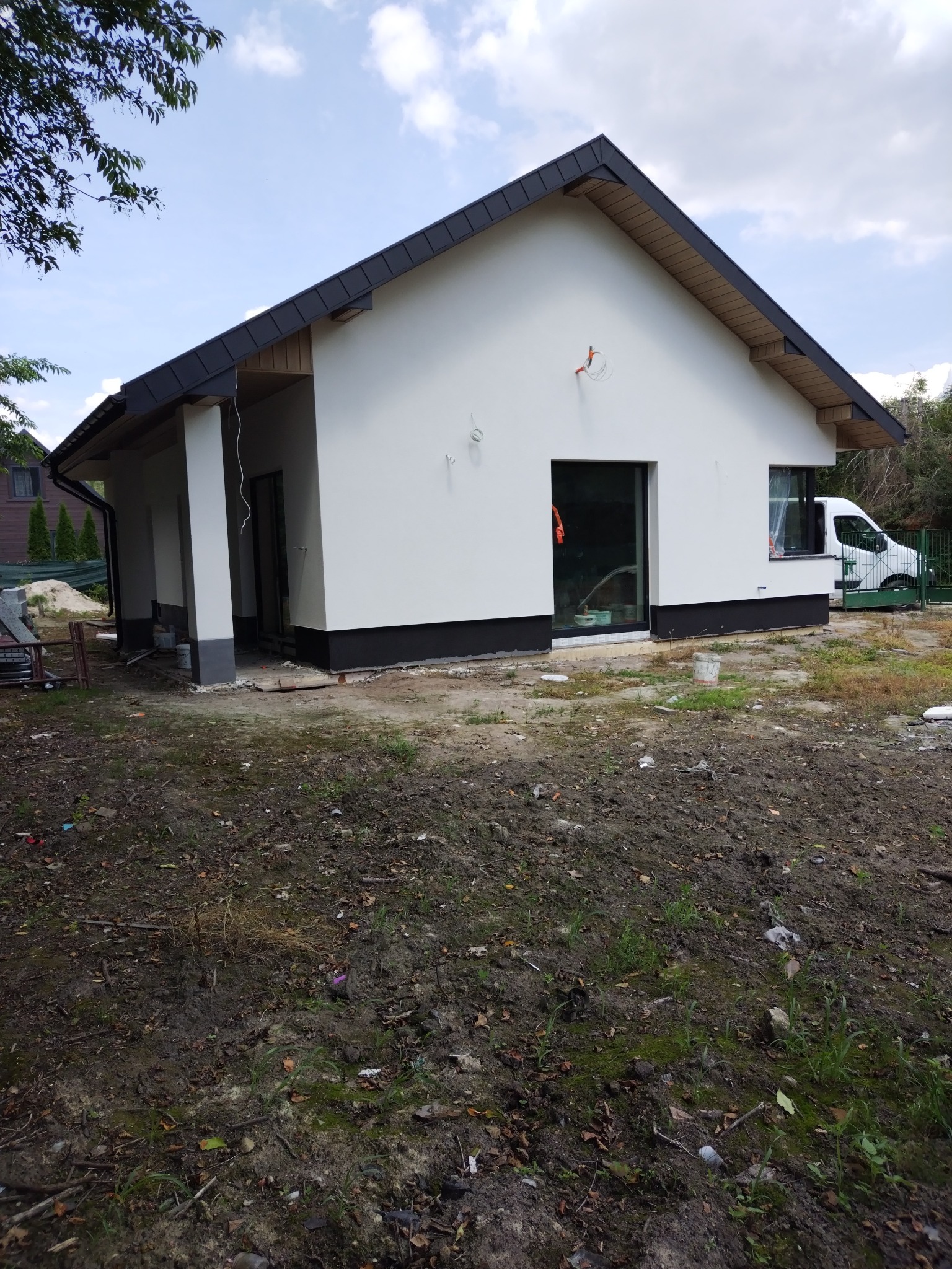 Niewielki, parterowy dom z białymi ścianami i ciemnym dachem, w trakcie wykańczania, widoczny na tle zieleni i błękitnego nieba z chmurami.