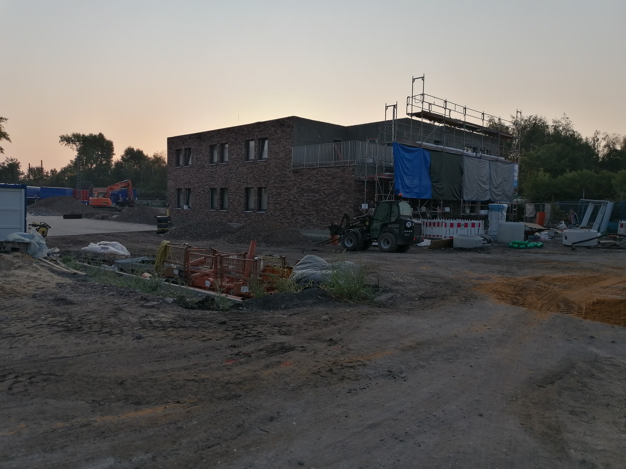 Dwupiętrowy budynek z cegły w trakcie budowy, otoczony rusztowaniami i sprzętem budowlanym, widziany o zmierzchu. Widoczny wykop i maszyny budowlane.