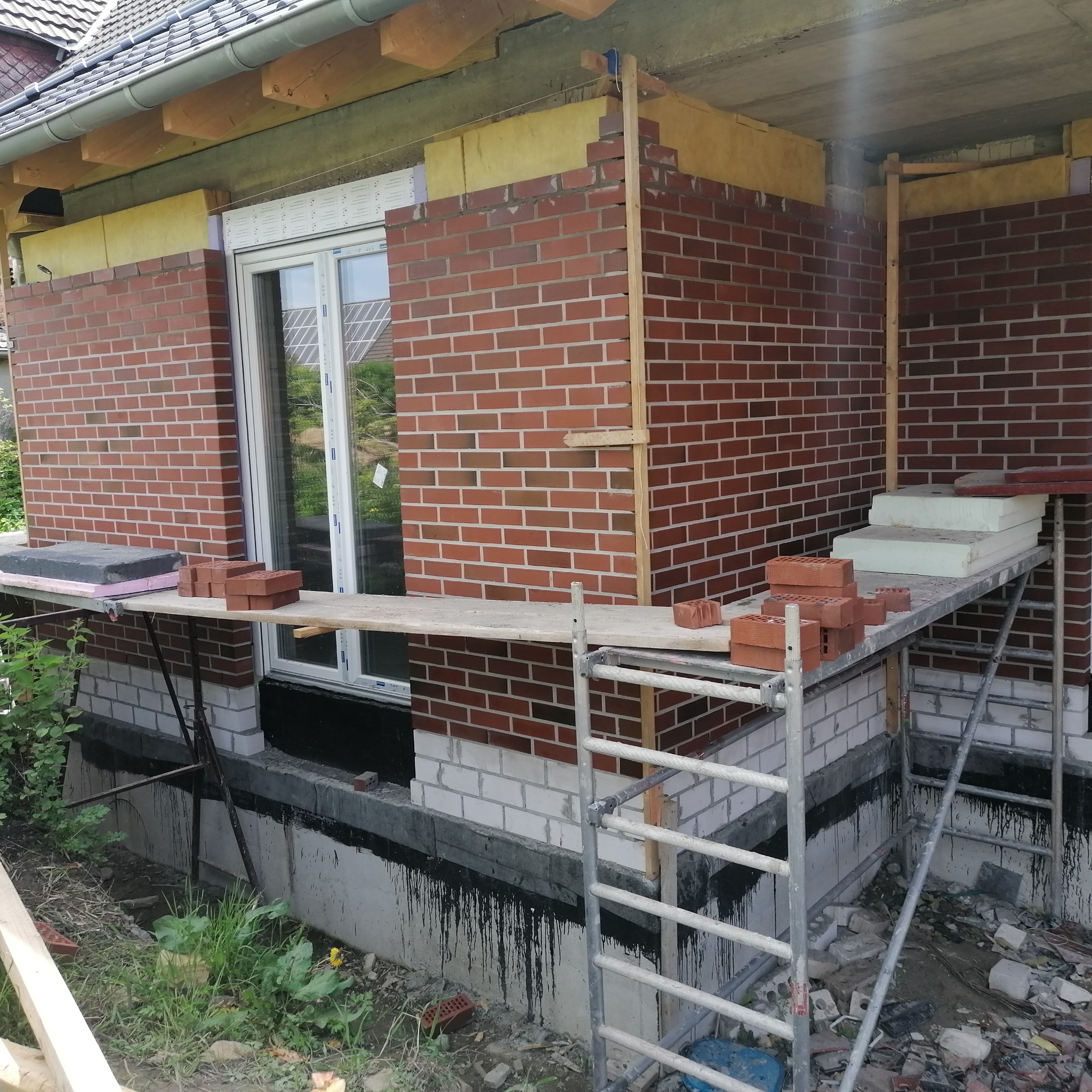 Murowanie ściany z czerwonej cegły, widoczne rusztowanie i zapas cegieł. W tle okno z odbiciem nieba. Etap budowy domu jednorodzinnego.