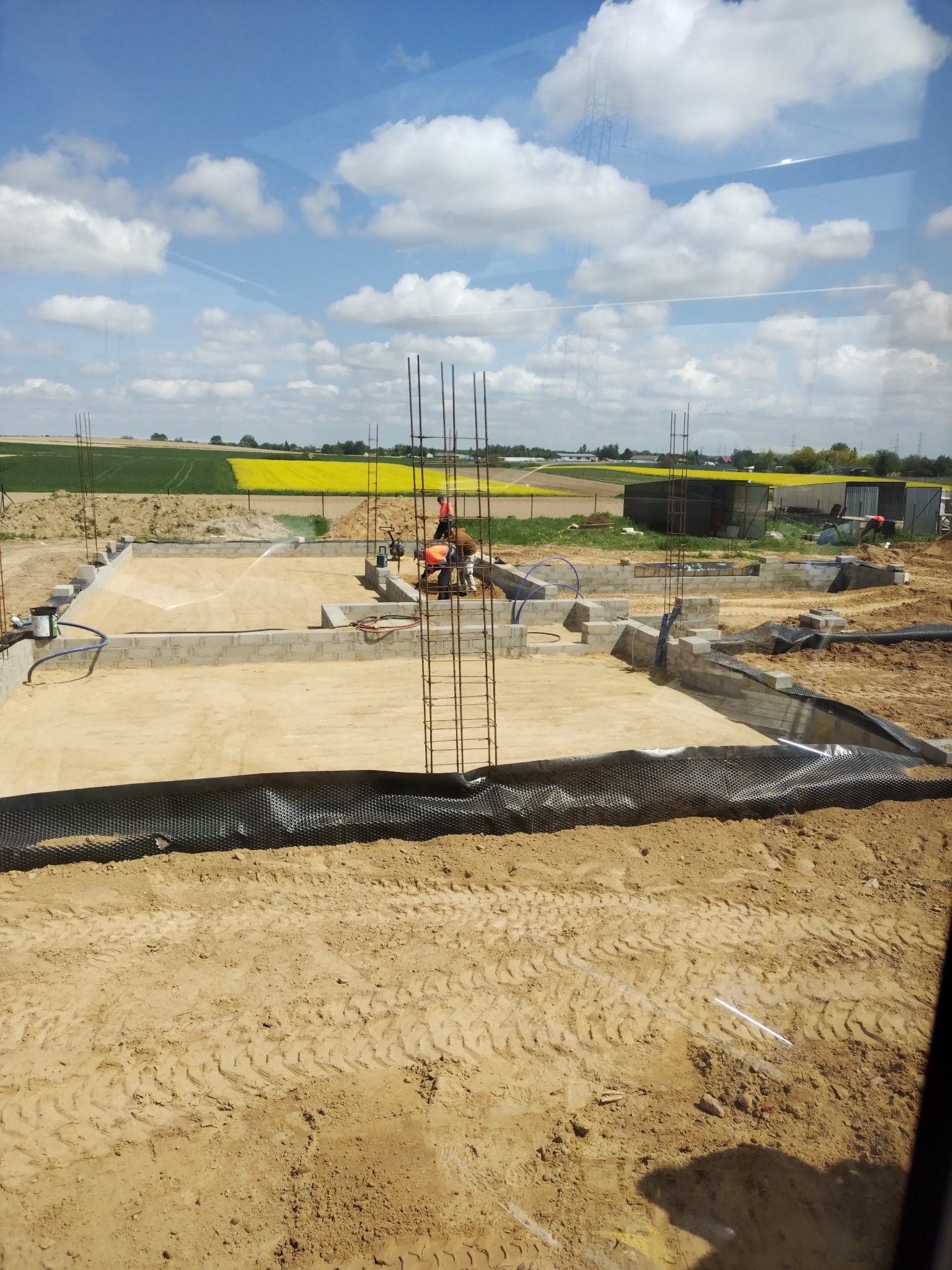 Fundament domu z bloczków betonowych, zbrojenie pionowe, folia fundamentowa. W tle pola uprawne i błękitne niebo z chmurami. Etap budowy w słoneczny dzień.