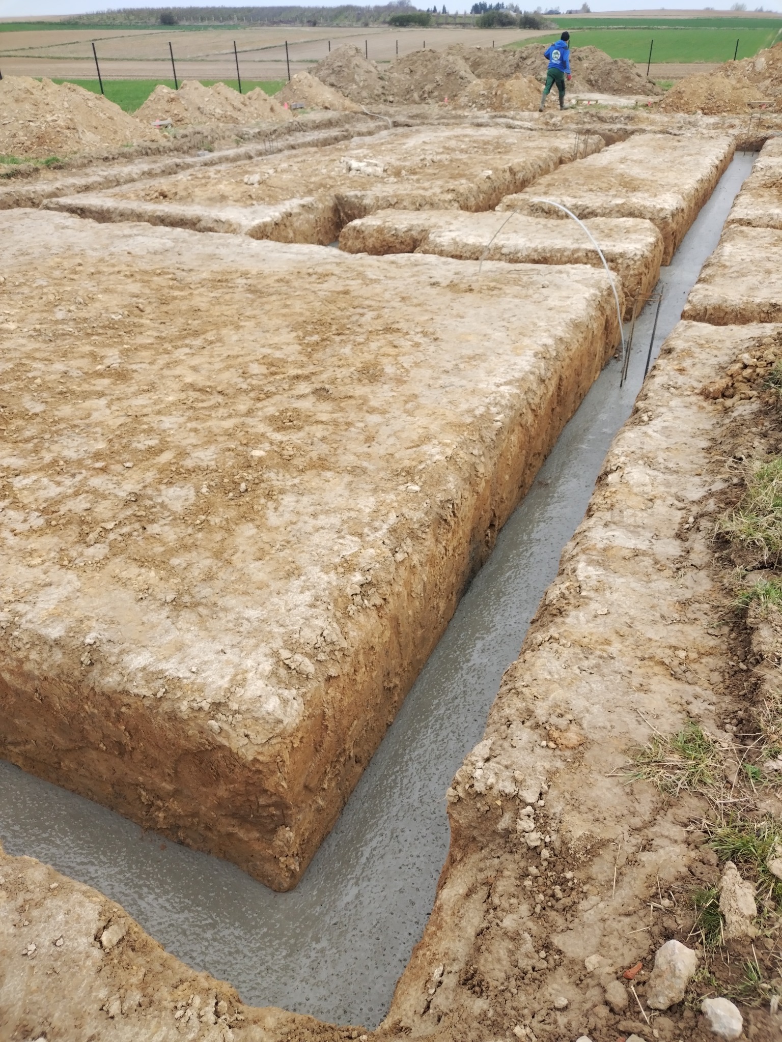 Wykopane i zalane betonem fundamenty pod budowę domu. Widoczne zbrojenia i kształt wykopu. W tle pole i osoba w oddali.