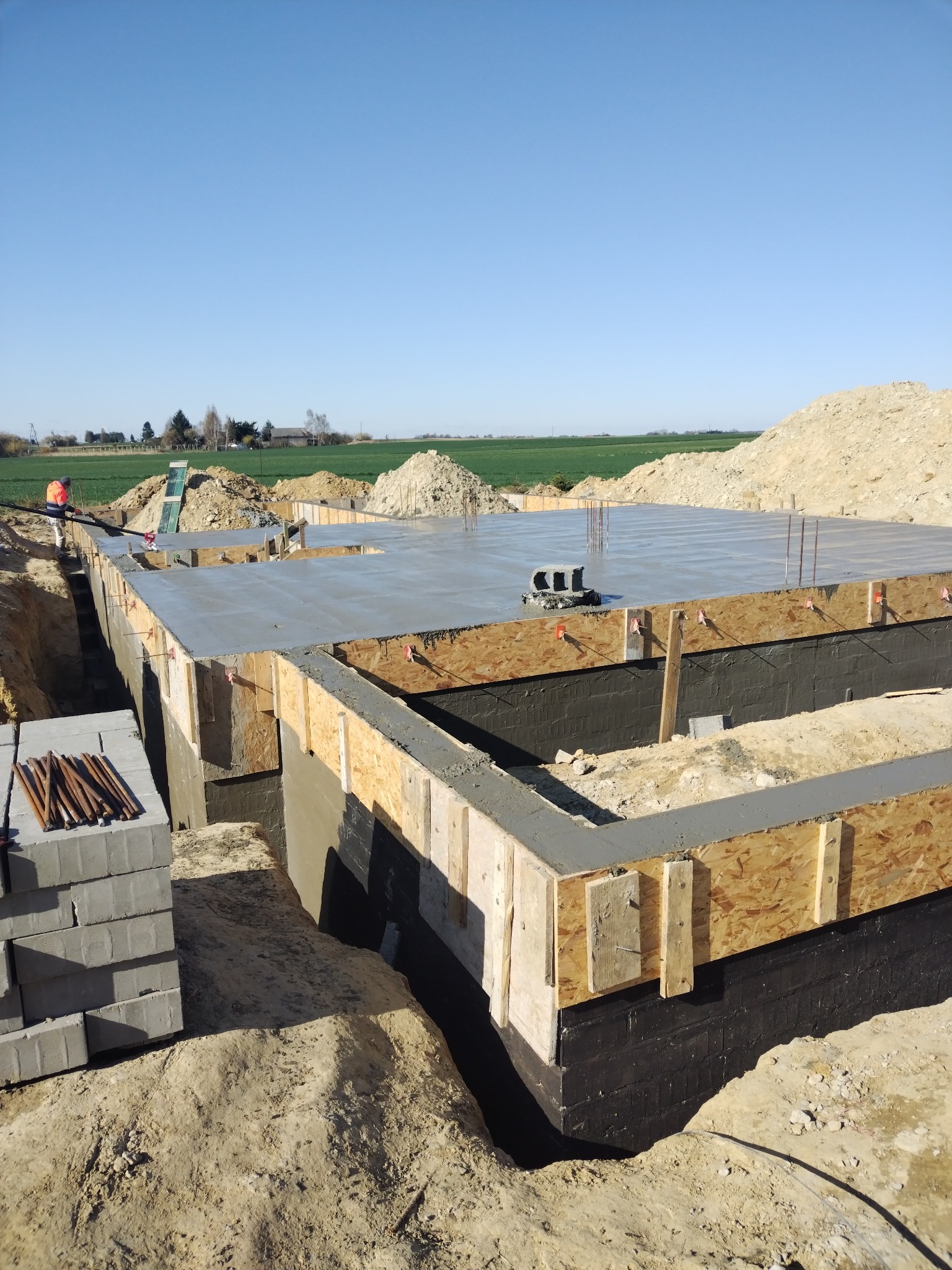 Zalane fundamenty domu z izolacją bitumiczną i szalunkiem drewnianym, widoczne zbrojenie, teren budowy z ziemią i zielonym polem w tle.