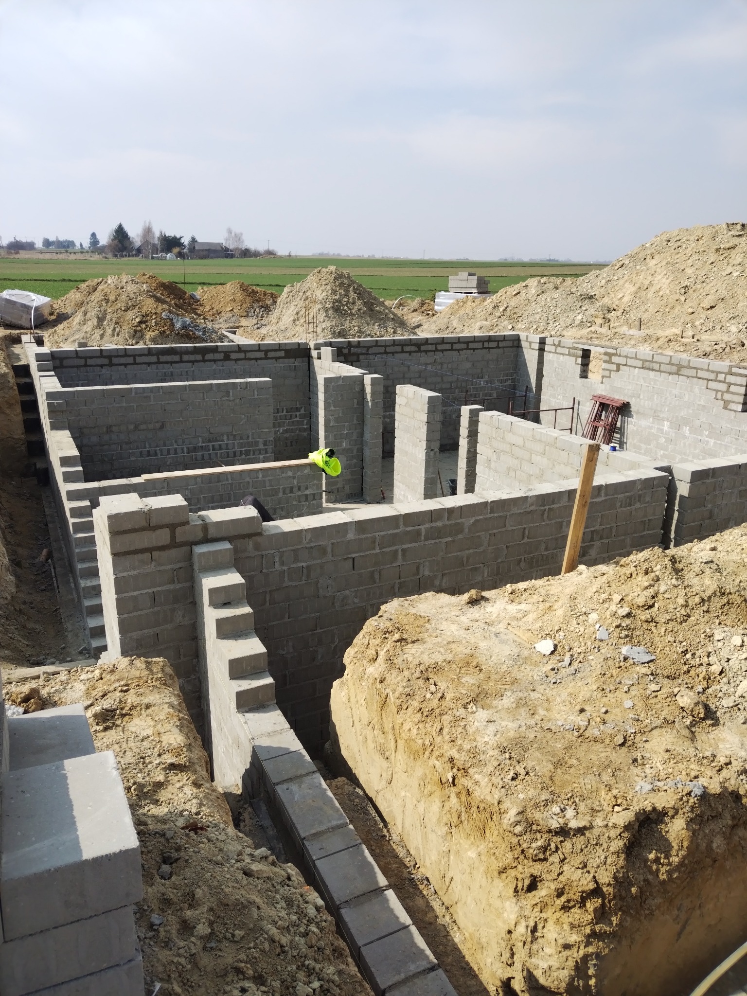 Wznoszenie fundamentów domu z bloczków betonowych na tle krajobrazu polnych. Widoczne wykopane rowy i ułożone bloczki. Żółta kamizelka na murze.