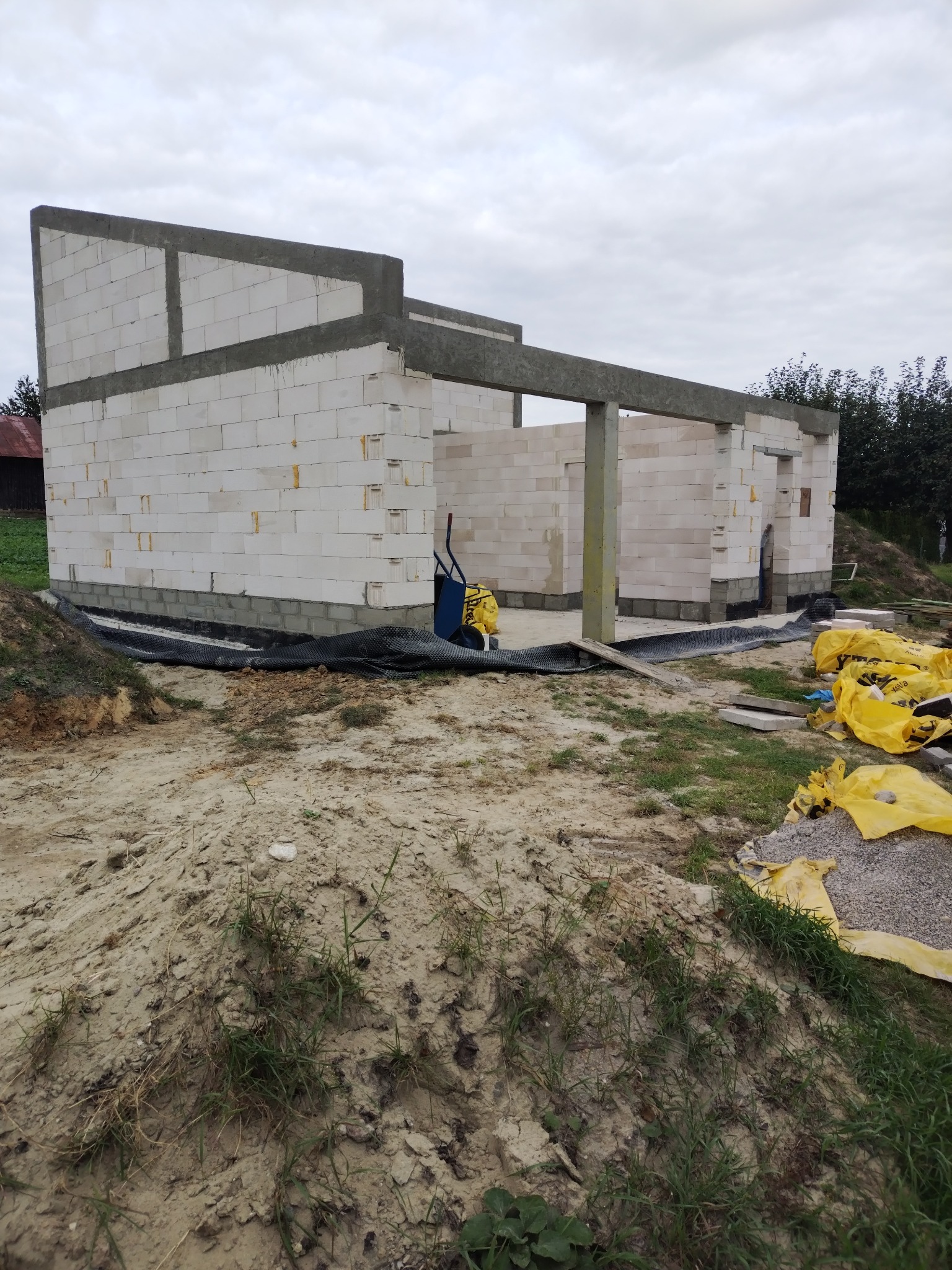 Wznoszona konstrukcja domu z białych bloczków, widoczne zbrojenia, częściowo wykonane ściany, teren budowy z workami materiałów budowlanych.