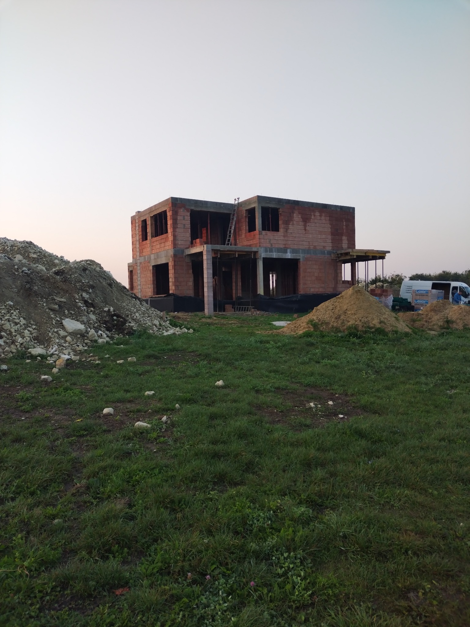 Surowy stan dwukondygnacyjnego domu z cegły, z widocznym szkieletem i stertami ziemi, na tle zielonej trawy i bladego nieba o zmierzchu. Widoczny częściowo samochód dostawczy.