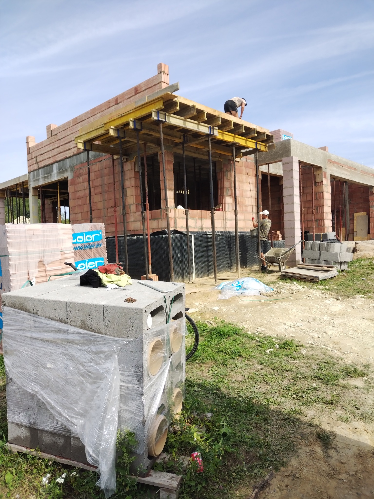 Budowa domu z czerwonej cegły: widoczne ściany, drewniany szalunek stropu i pracujący na górze robotnik. Na pierwszym planie bloczki betonowe w folii.