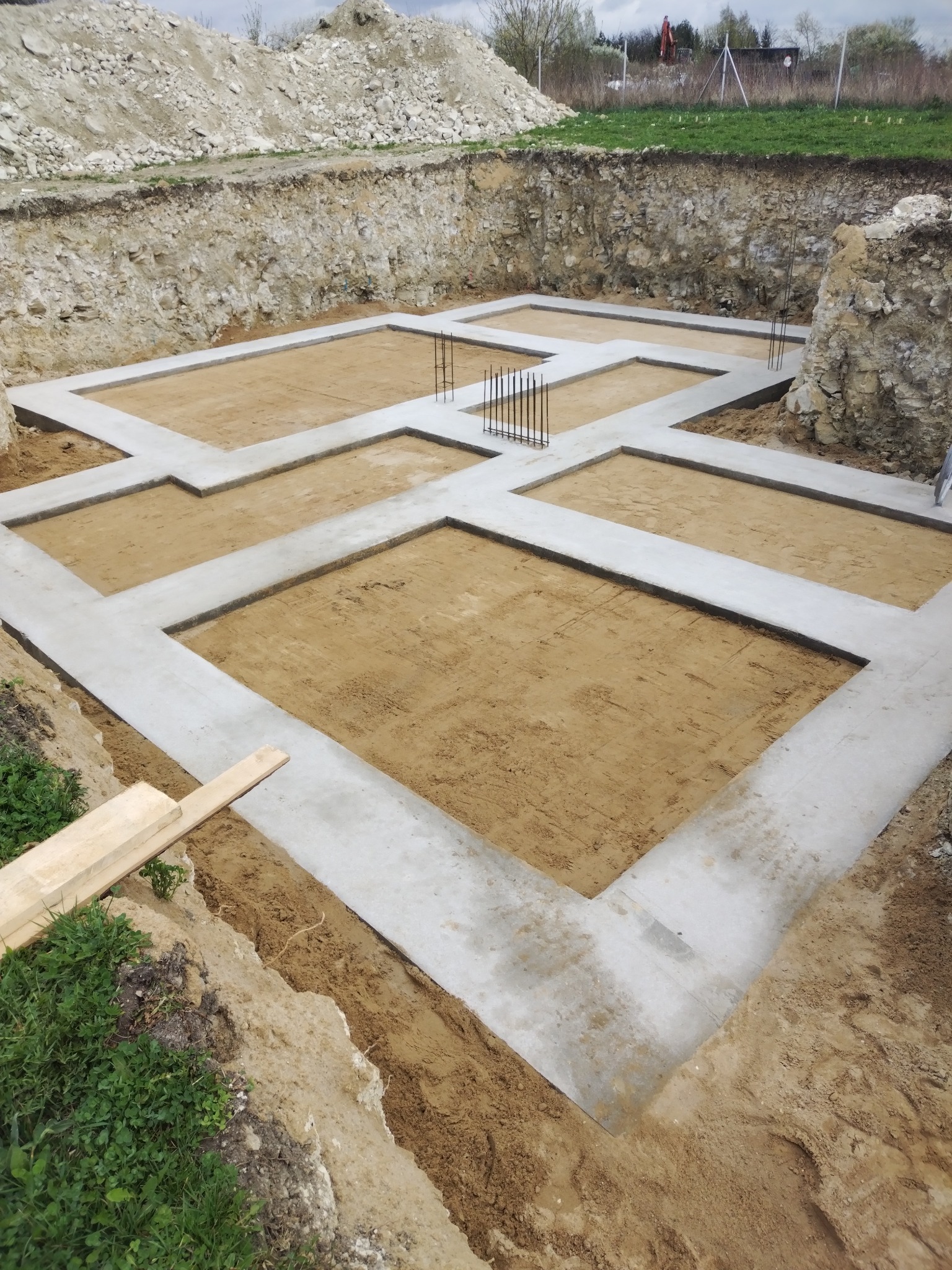 Zalane fundamenty pod budynek z wystającymi prętami zbrojeniowymi, widoczne w wykopie. Ujęcie z góry, w tle ziemia i zieleń.