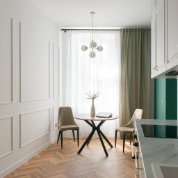 Mar-Mur Mariusz Mnochy - Jasne wnętrze: jadalnia z okrągłym stołem na tle okna z zasłonami, parkiet w jodełkę, dekoracyjne listwy na ścianie, widok na fragment blatu kuchennego i szafki.