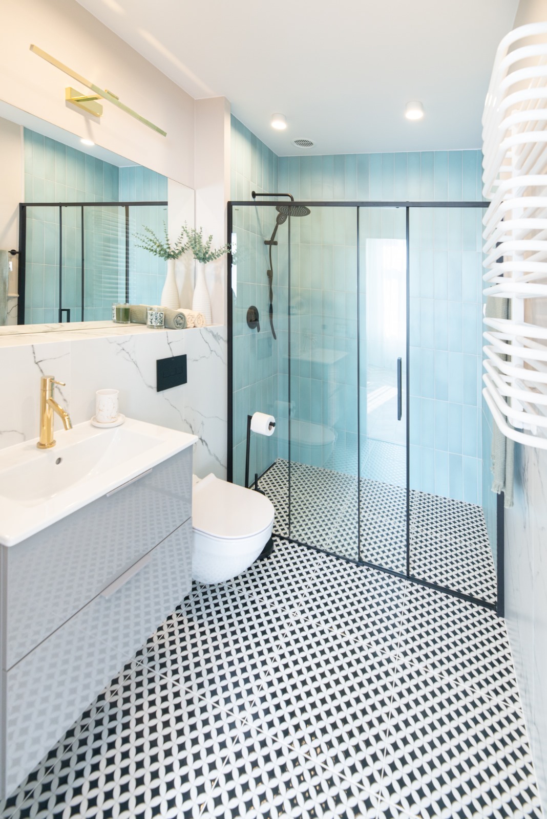 Nowoczesna łazienka z kabiną prysznicową typu walk-in, umywalką nablatową z baterią w kolorze złota, białą toaletą i geometryczną posadzką w czarno-białe wzory. Ściany wyłożone turkusowymi płytkami.