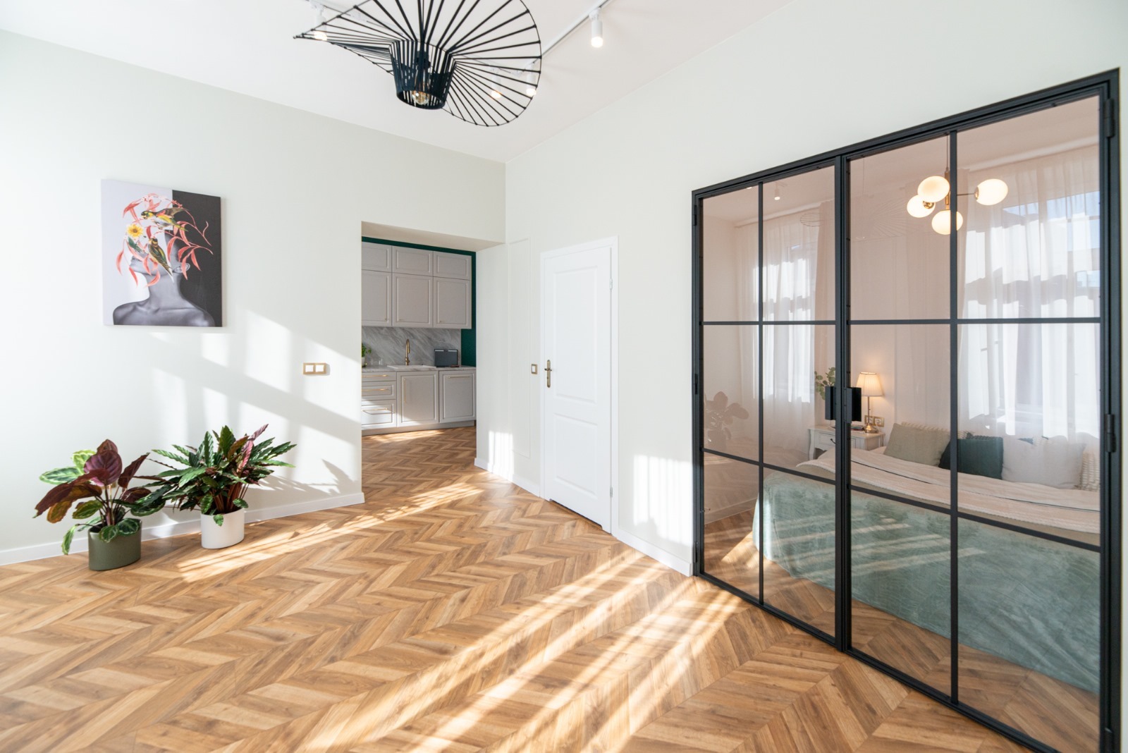 Jasne wnętrze z parkietem w jodełkę, czarną ścianą działową ze szkła, widok na kuchnię i sypialnię, ozdobione roślinami doniczkowymi i abstrakcyjnym obrazem.
