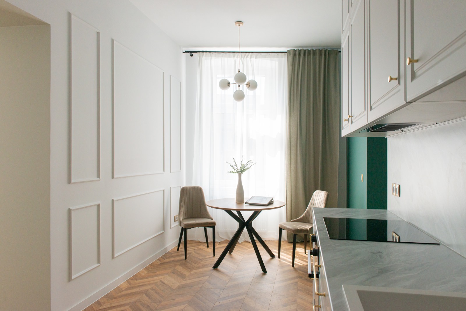 Jasne wnętrze: jadalnia z okrągłym stołem na tle okna z zasłonami, parkiet w jodełkę, dekoracyjne listwy na ścianie, widok na fragment blatu kuchennego i szafki.