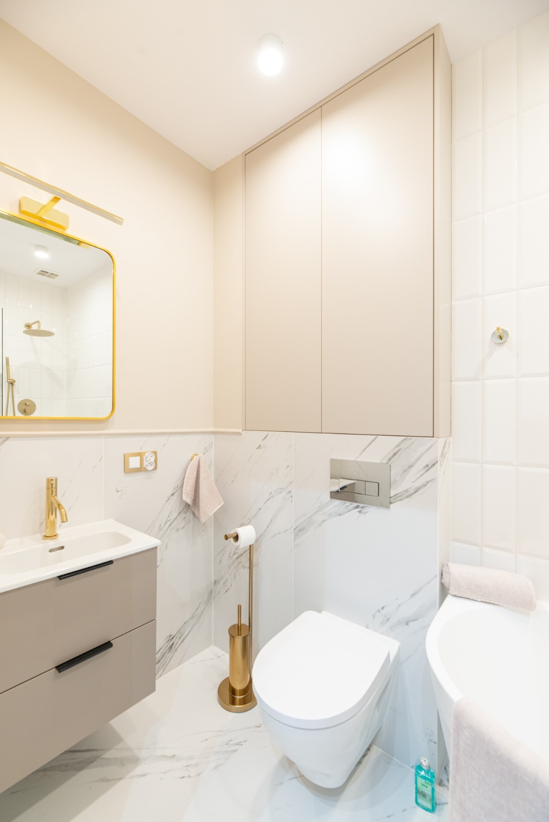 Jasna łazienka z marmurowymi płytkami, złotą armaturą i akcesoriami, wiszącą toaletą oraz szafką nad zabudową WC.