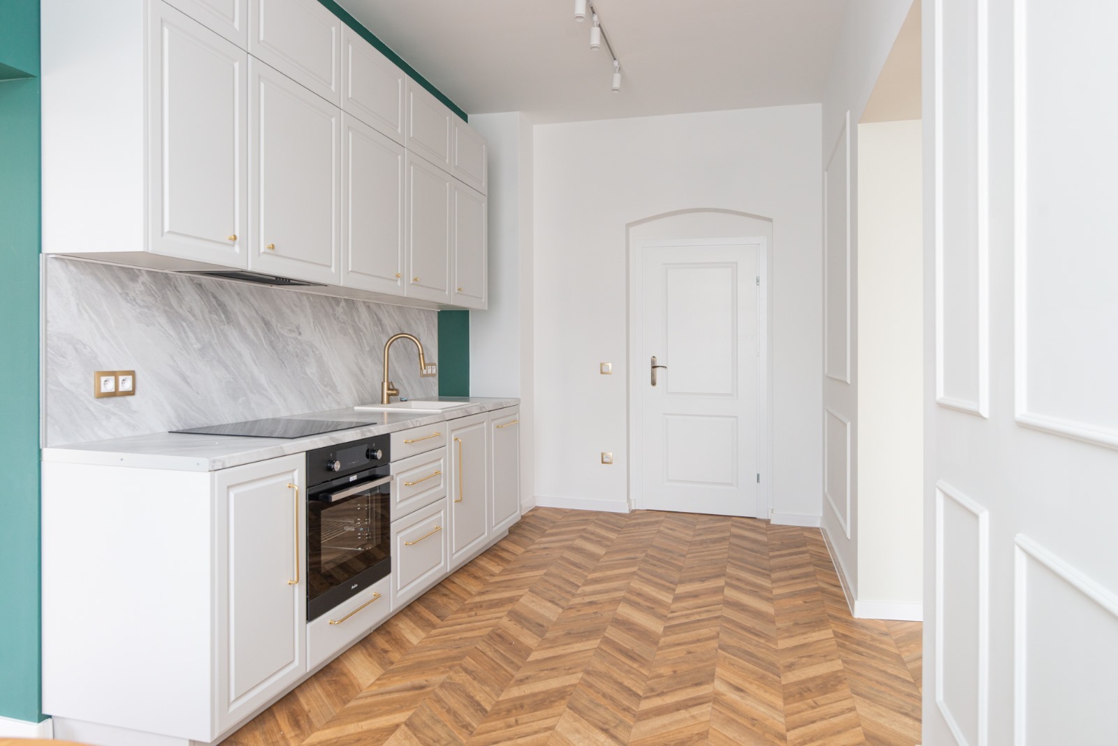 Jasna kuchnia z białymi szafkami, marmurowym backsplashiem i złotymi detalami, parkiet w jodełkę, widok na białe drzwi i ozdobne panele ścienne.