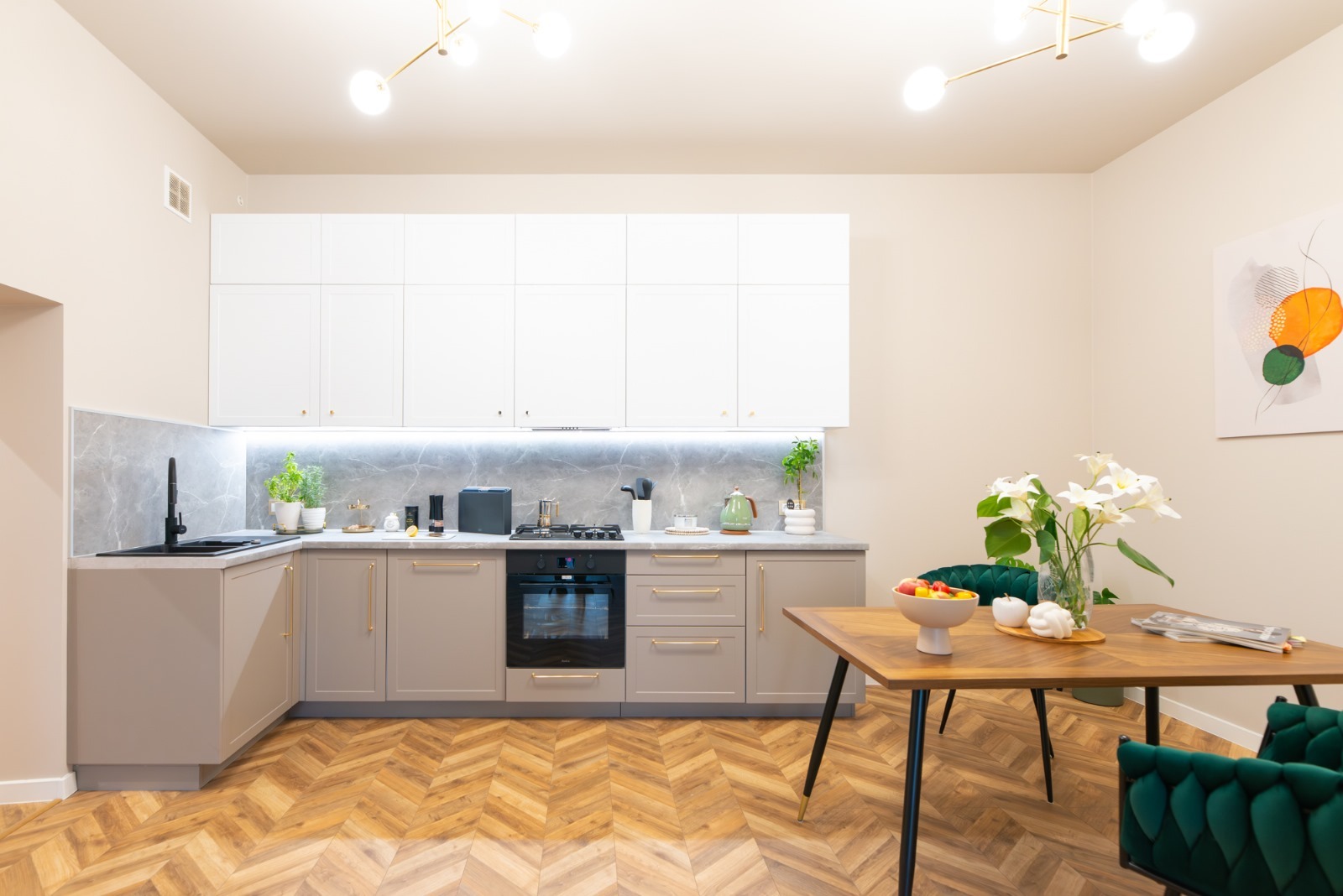 Jasna kuchnia z drewnianą podłogą w jodełkę, szarymi szafkami dolnymi z złotymi uchwytami i białymi górnymi, marmurowym backsplashiem, czarnym zlewem i baterią, oraz stołem z owocami i kwiatami...