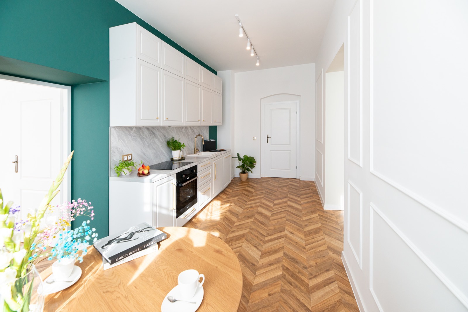 Nowoczesna kuchnia z białymi szafkami, marmurowym blatem, parkietem w jodełkę i akcentem turkusowej ściany, oświetlona światłem słonecznym.