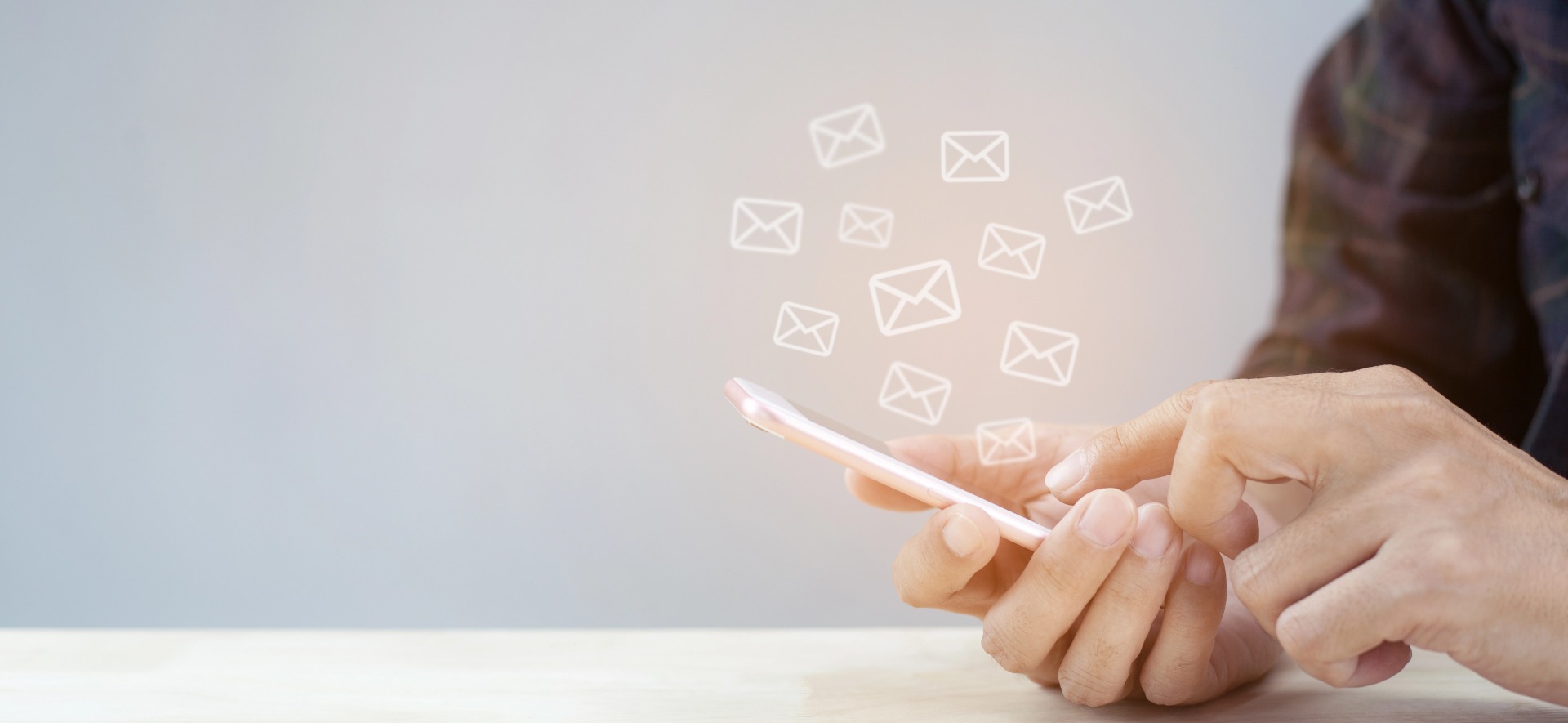 Ręka mężczyzny obsługuje smartfon z ikonami kopert, symbolizującymi wiadomości e-mail, na tle szarej ściany.