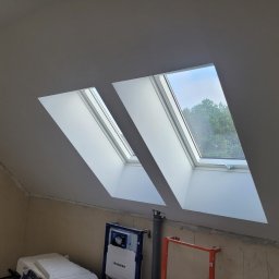 remoglass9 - Dwa okna dachowe w szarej ścianie poddasza, widoczne drzewa za oknem, poniżej instalacja sanitarna Geberit i fragment rusztowania.