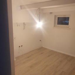 remoglass9 - Wnętrze pokoju w trakcie remontu: położona podłoga z paneli, ściany przygotowane do szpachlowania, widoczne kable elektryczne, surowy sufit z belkami i wiszącą żarówką.