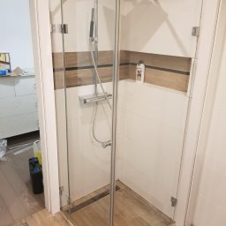 remoglass9 - Nowoczesna kabina prysznicowa ze szklanymi ścianami, chromowanymi okuciami i deszczownicą, w łazience z białymi płytkami i drewnopodobnymi akcentami, widoczna butelka kosmetyku w niszy.