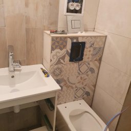 remoglass9 - Montaż podtynkowej instalacji sanitarnej w łazience z widoczną zabudową WC w mozaikę, umywalką z szafką i otwartą skrzynką z wodomierzami.