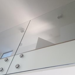 remoglass9 - Minimalistyczna szklana balustrada wewnętrzna z metalowymi mocowaniami punktowymi, widok z dołu na sufit z oświetleniem.