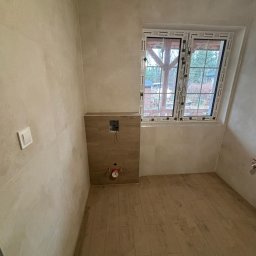 remoglass9 - Surowe wnętrze łazienki w trakcie remontu: beżowe płytki na ścianach i podłodze imitujące drewno, przygotowane miejsce na stelaż podtynkowy WC, okno z widokiem na ogród.