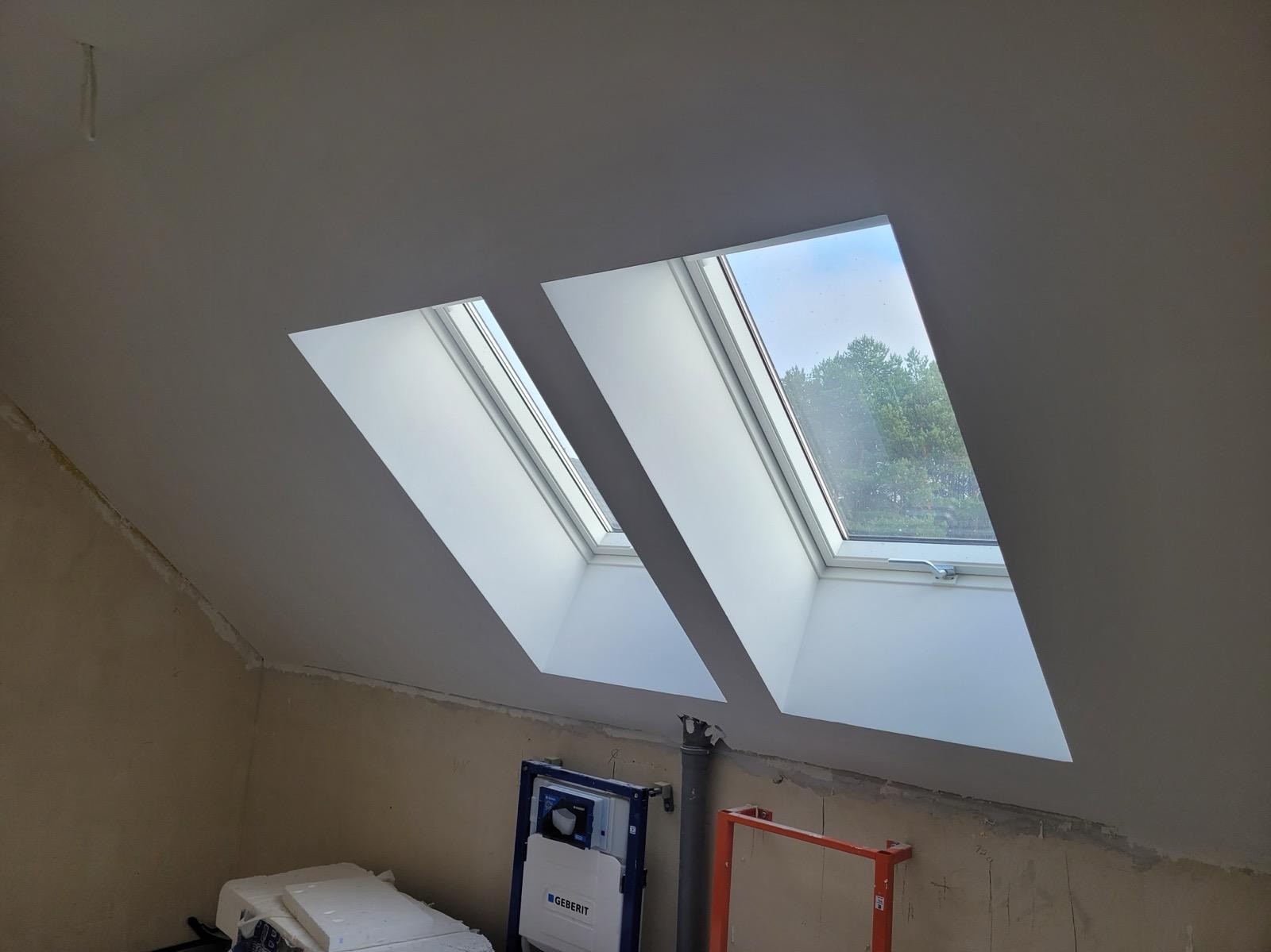 Dwa okna dachowe w szarej ścianie poddasza, widoczne drzewa za oknem, poniżej instalacja sanitarna Geberit i fragment rusztowania.