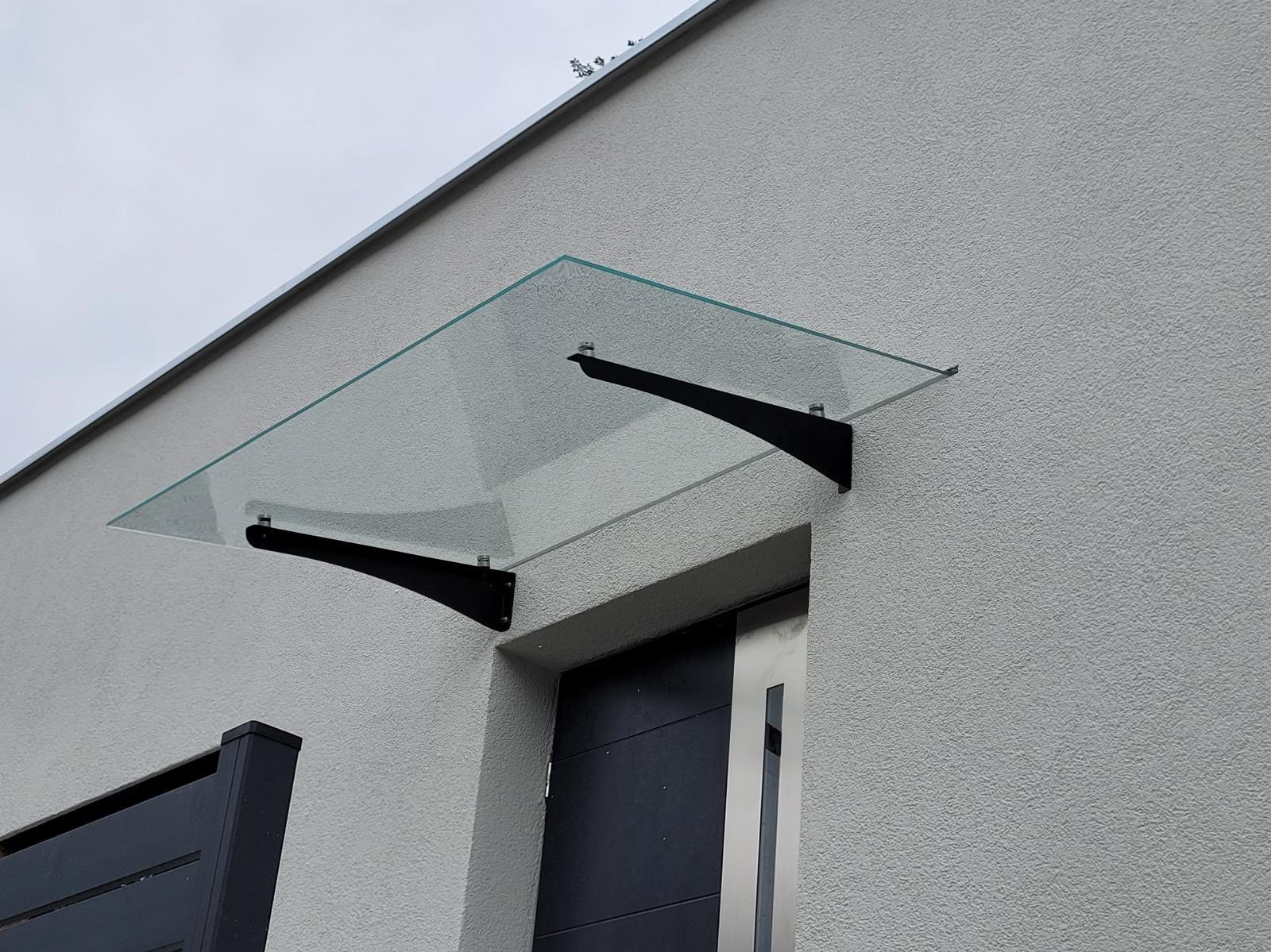 Szklany daszek nad wejściem, wsparty na dwóch czarnych, metalowych wspornikach, przymocowany do ściany o szarej, chropowatej fakturze.