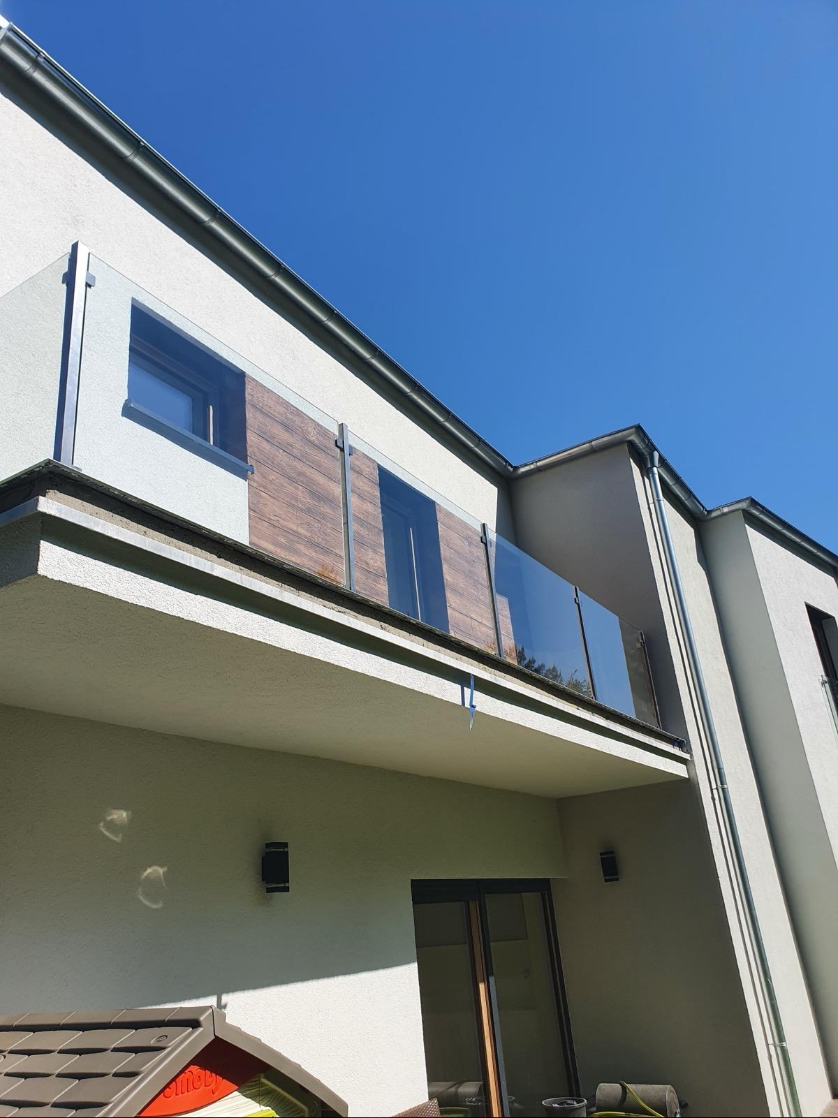 Nowoczesny balkon z szklaną balustradą, fragmentami wykończonymi okładziną imitującą drewno, widok z dołu na tle błękitnego nieba.