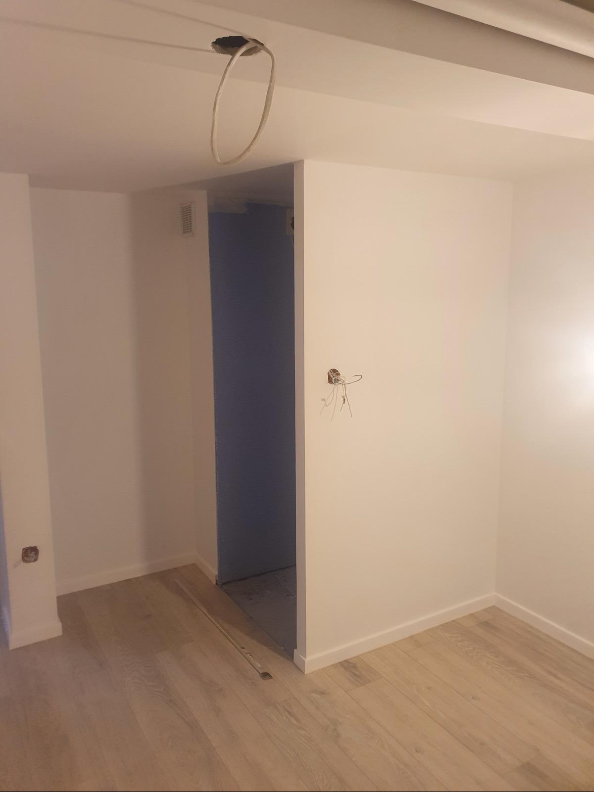 Wnętrze w trakcie remontu: białe ściany, drewniana podłoga, widoczne kable wystające ze ściany, otwór w suficie z kablami, surowy stan przed wykończeniem.