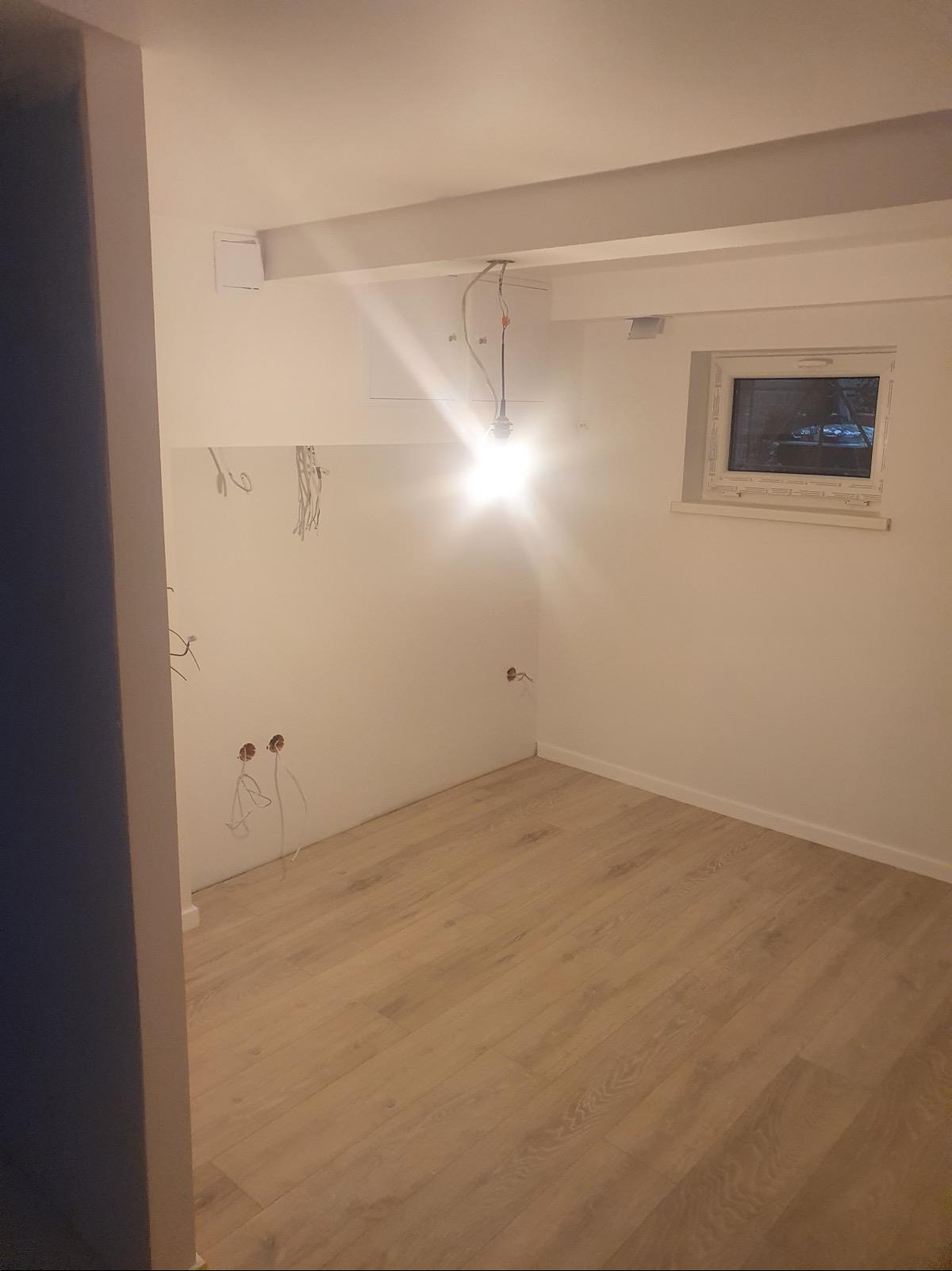 Wnętrze pokoju w trakcie remontu: położona podłoga z paneli, ściany przygotowane do szpachlowania, widoczne kable elektryczne, surowy sufit z belkami i wiszącą żarówką.