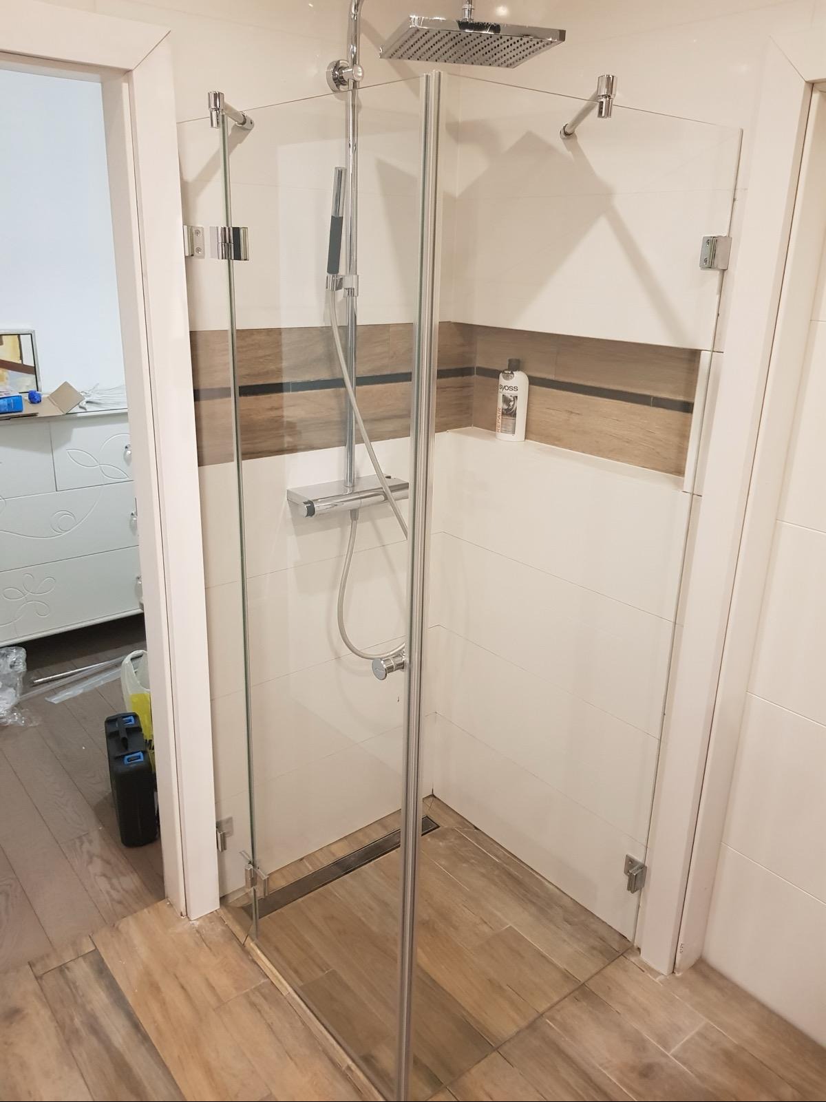 Nowoczesna kabina prysznicowa ze szklanymi ścianami, chromowanymi okuciami i deszczownicą, w łazience z białymi płytkami i drewnopodobnymi akcentami, widoczna butelka kosmetyku w niszy.