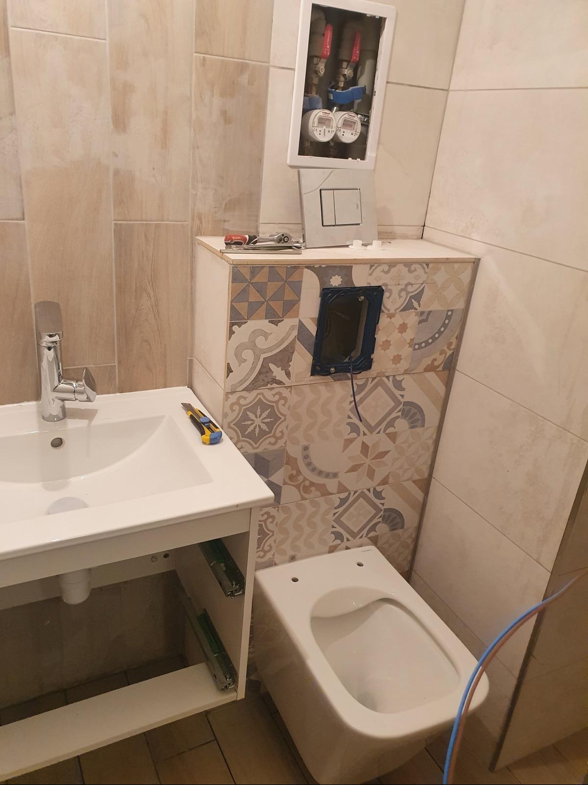 Montaż podtynkowej instalacji sanitarnej w łazience z widoczną zabudową WC w mozaikę, umywalką z szafką i otwartą skrzynką z wodomierzami.