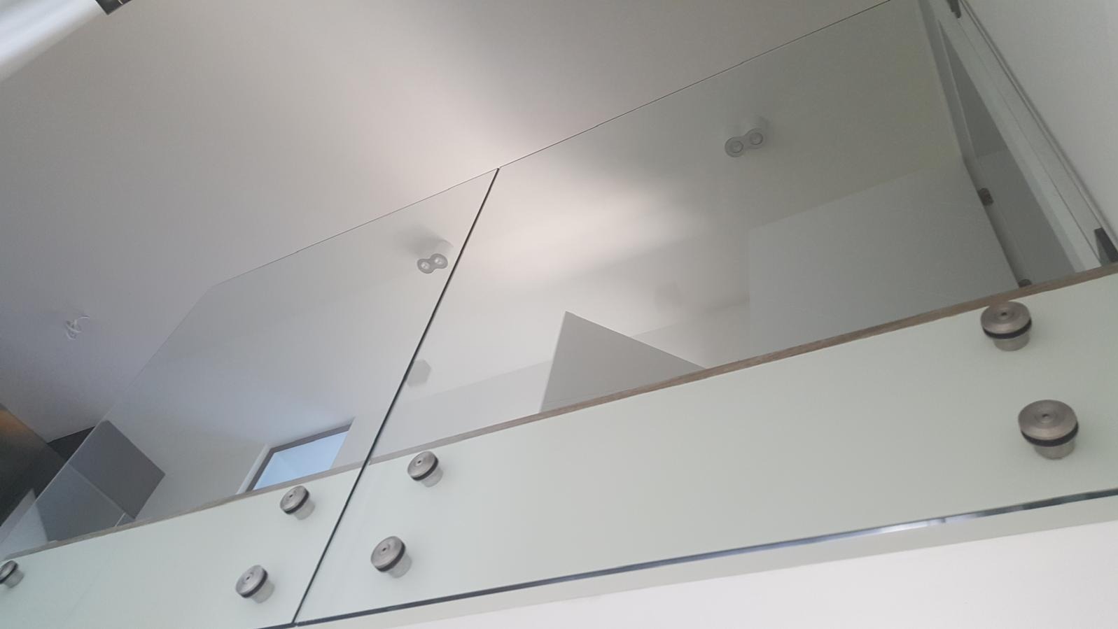 Minimalistyczna szklana balustrada wewnętrzna z metalowymi mocowaniami punktowymi, widok z dołu na sufit z oświetleniem.