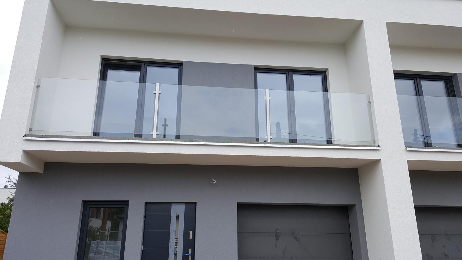 Nowoczesny balkon z balustradą ze szkła i stali nierdzewnej, widok z dołu na dwukondygnacyjny budynek z szarą fasadą i czarnymi ramami okiennymi.
