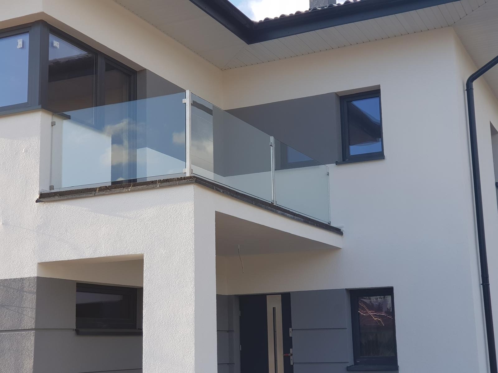 Nowoczesny balkon z przezroczystą szklaną balustradą, wspartą na srebrnych słupkach, odbijającą błękitne niebo i chmury, na tle białej elewacji budynku z szarymi akcentami.