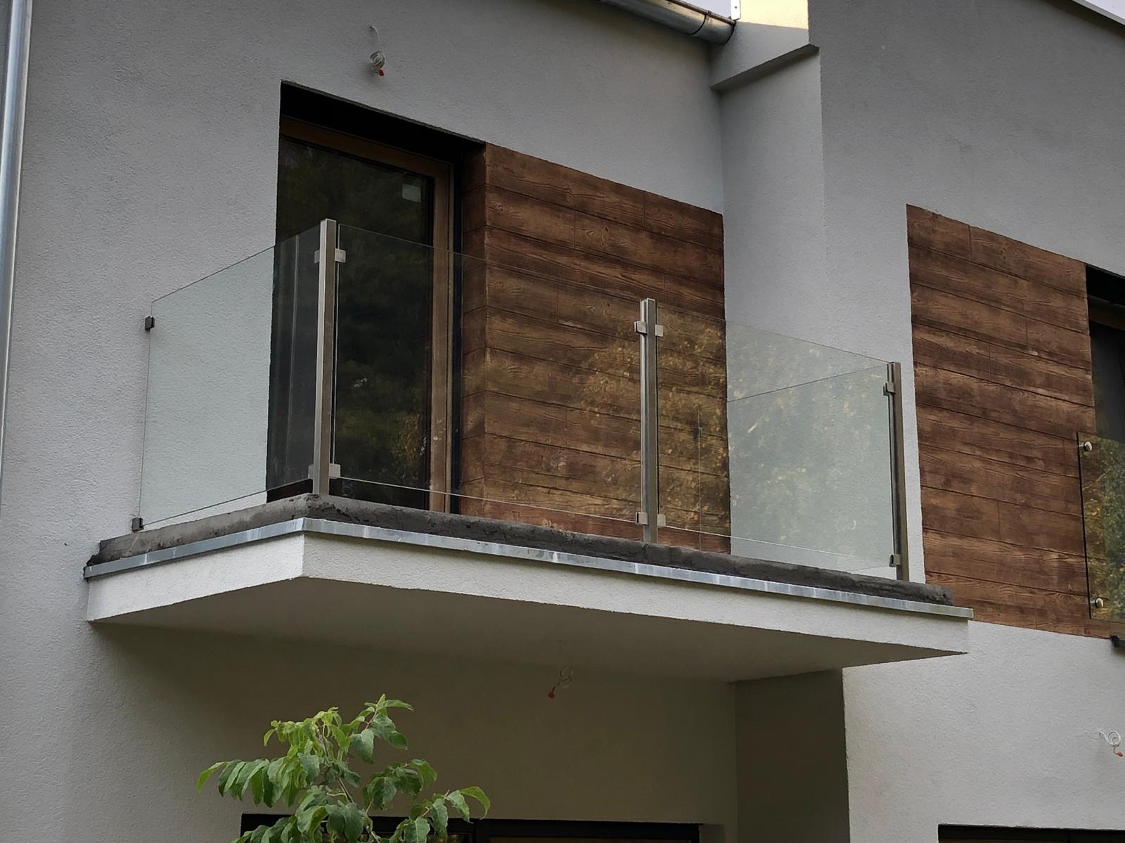 Nowoczesny balkon z szklaną balustradą na metalowych słupkach, z widoczną elewacją imitującą drewno i fragmentem zieleni.