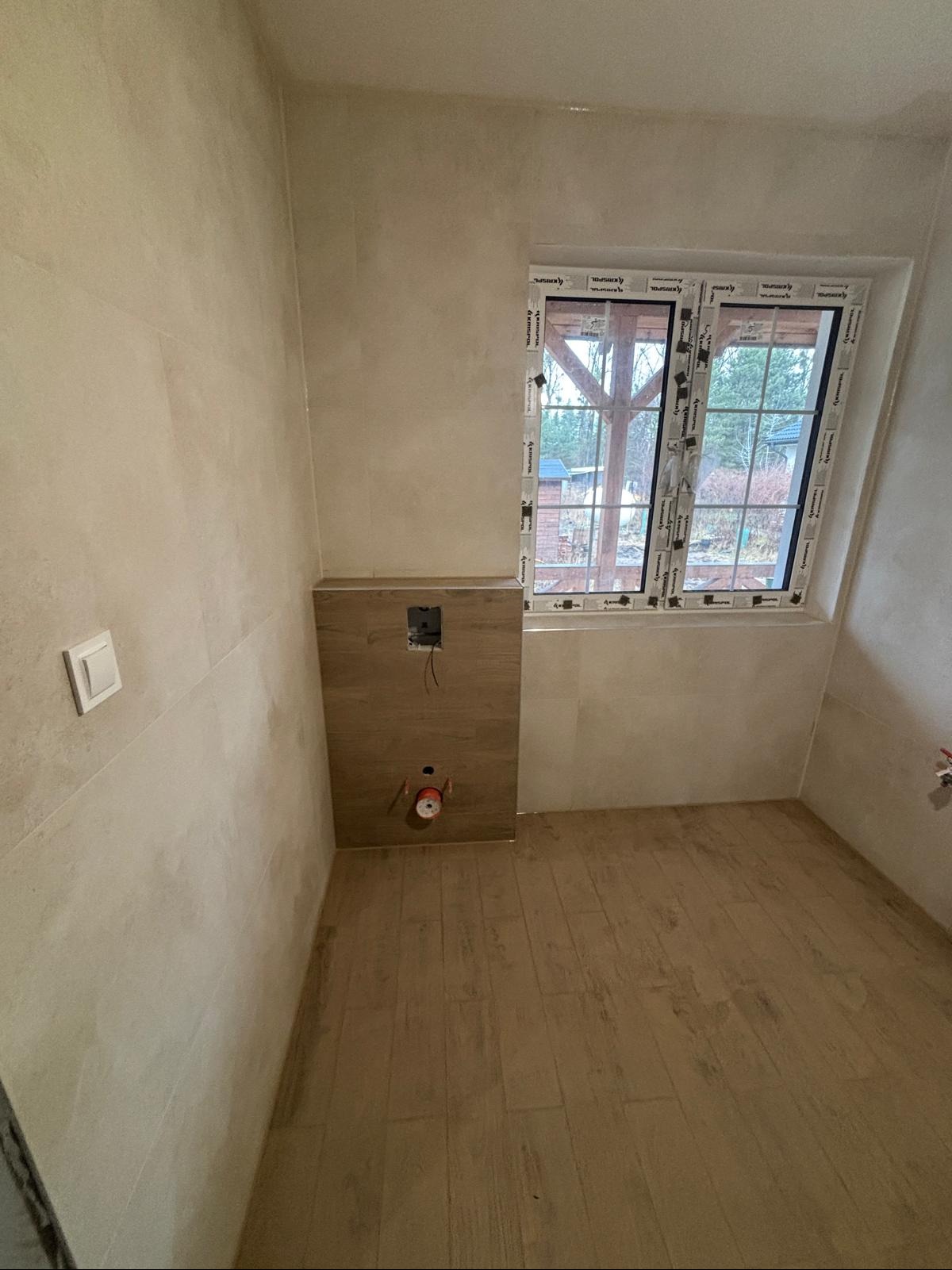 Surowe wnętrze łazienki w trakcie remontu: beżowe płytki na ścianach i podłodze imitujące drewno, przygotowane miejsce na stelaż podtynkowy WC, okno z widokiem na ogród.