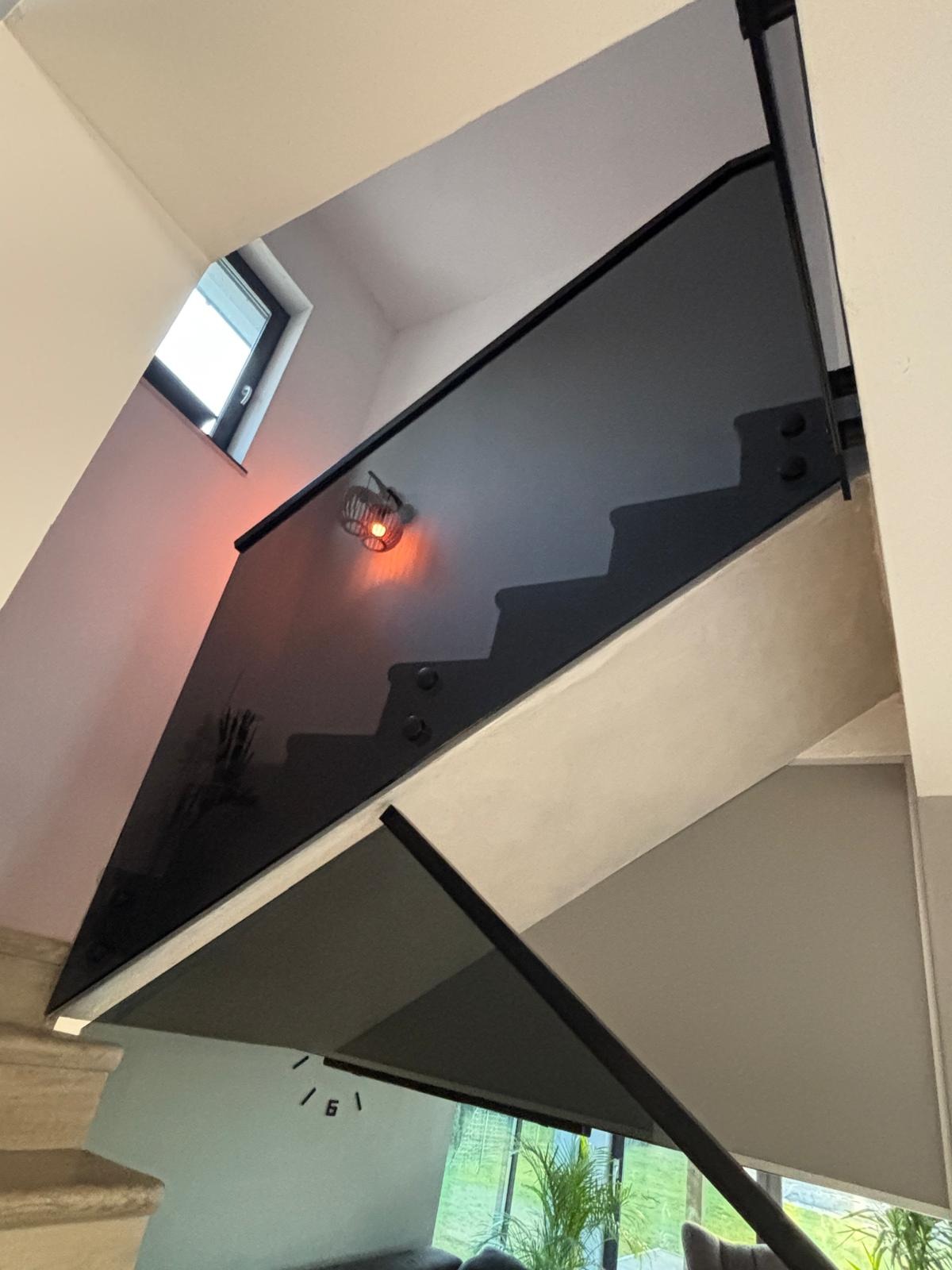 Szklana balustrada schodowa z czarnymi okuciami, widok z dołu, oświetlona lampą ścienną, odbicie schodów w szkle.