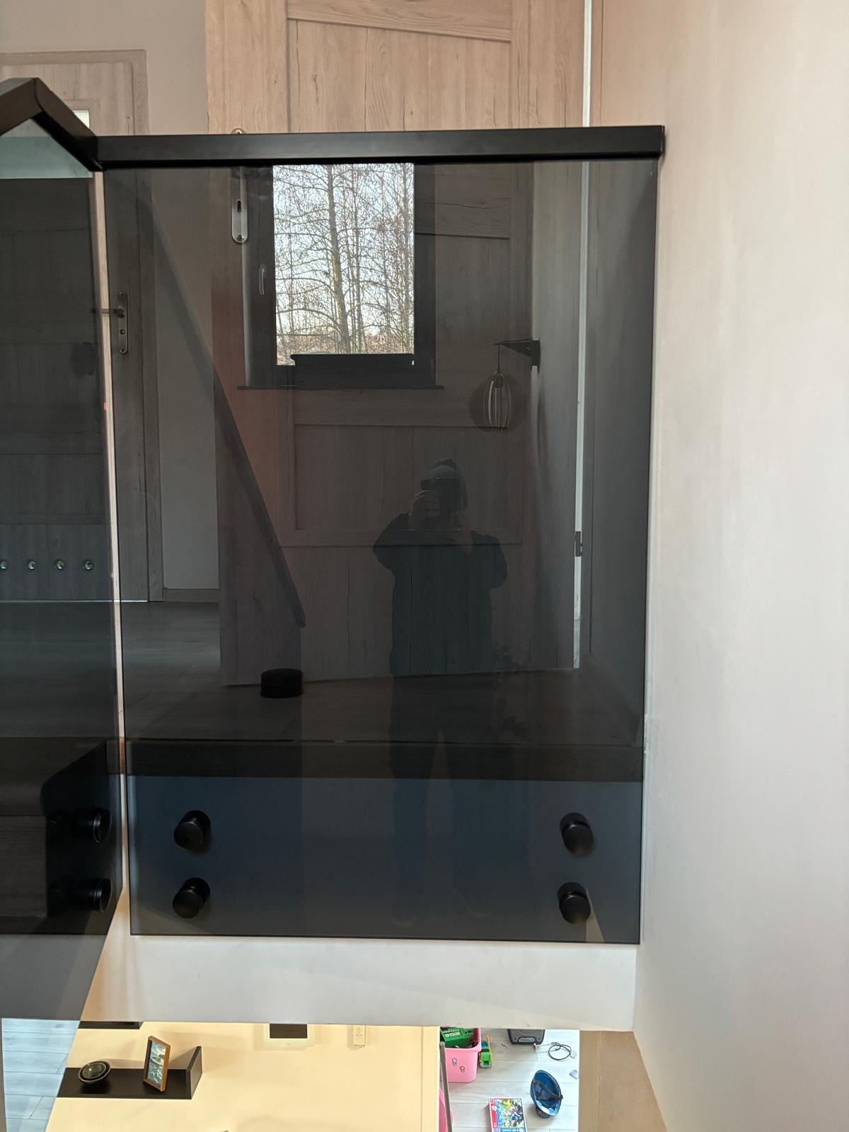 Szklana balustrada schodowa z czarnymi elementami mocującymi, widoczne odbicie fotografa w szkle.