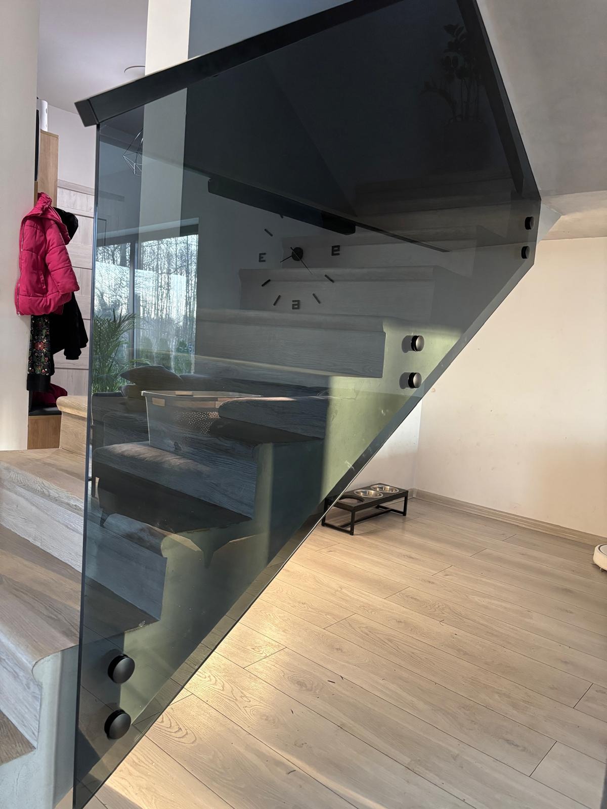 Nowoczesne schody z drewnianymi stopniami i szklaną balustradą z czarnymi mocowaniami, widok z dołu, na ścianie zegar, na podłodze miski dla zwierząt.