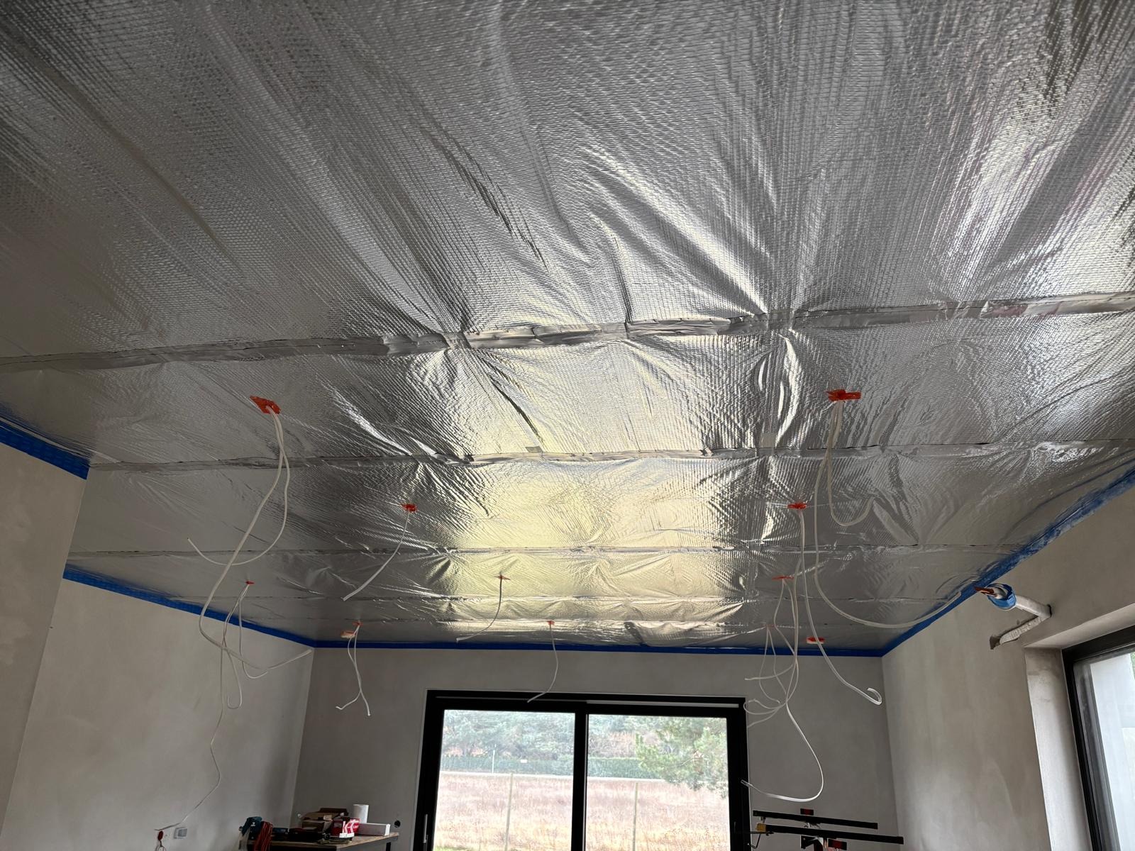 Izolacja termiczna sufitu folią aluminiową z wystającymi przewodami elektrycznymi w nowym budynku, widok z dołu na okno z widokiem na zewnątrz.