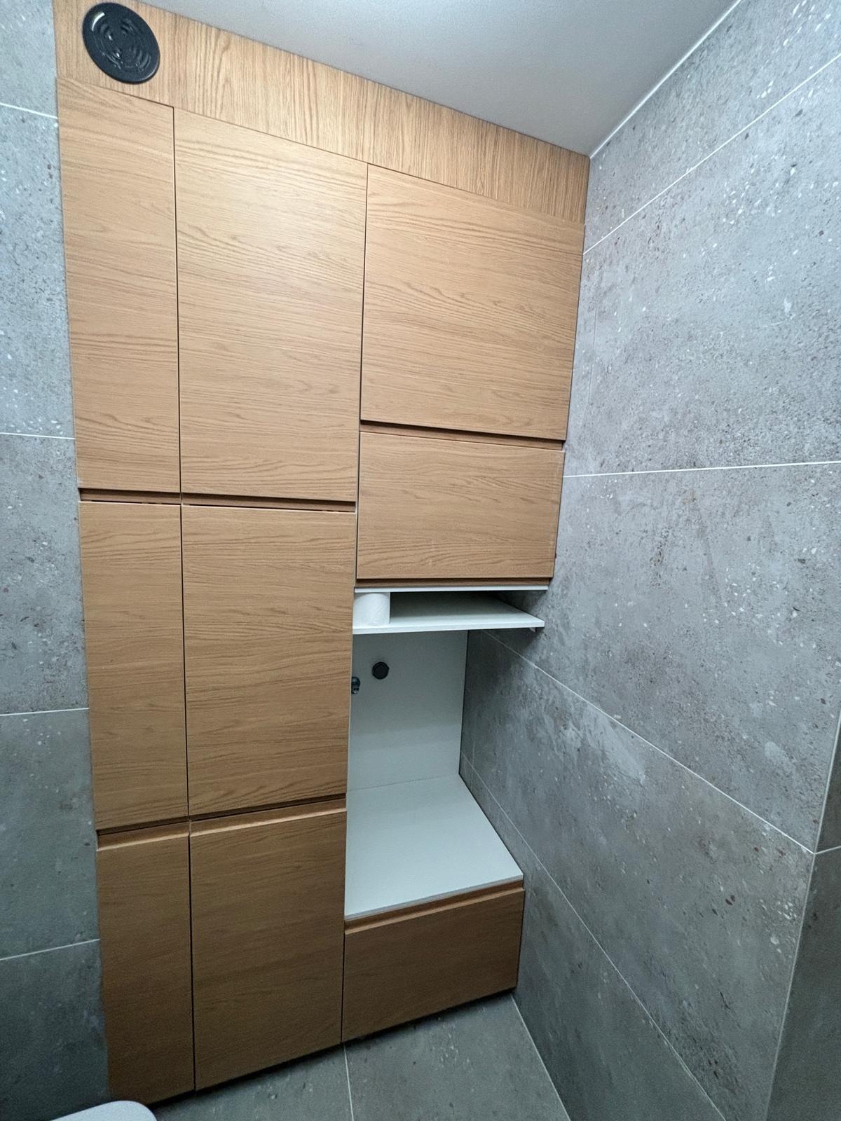 Drewniana zabudowa meblowa w łazience z szarą, kamienną okładziną ścienną. Widoczna biała półka i fragment instalacji. Minimalistyczny design.