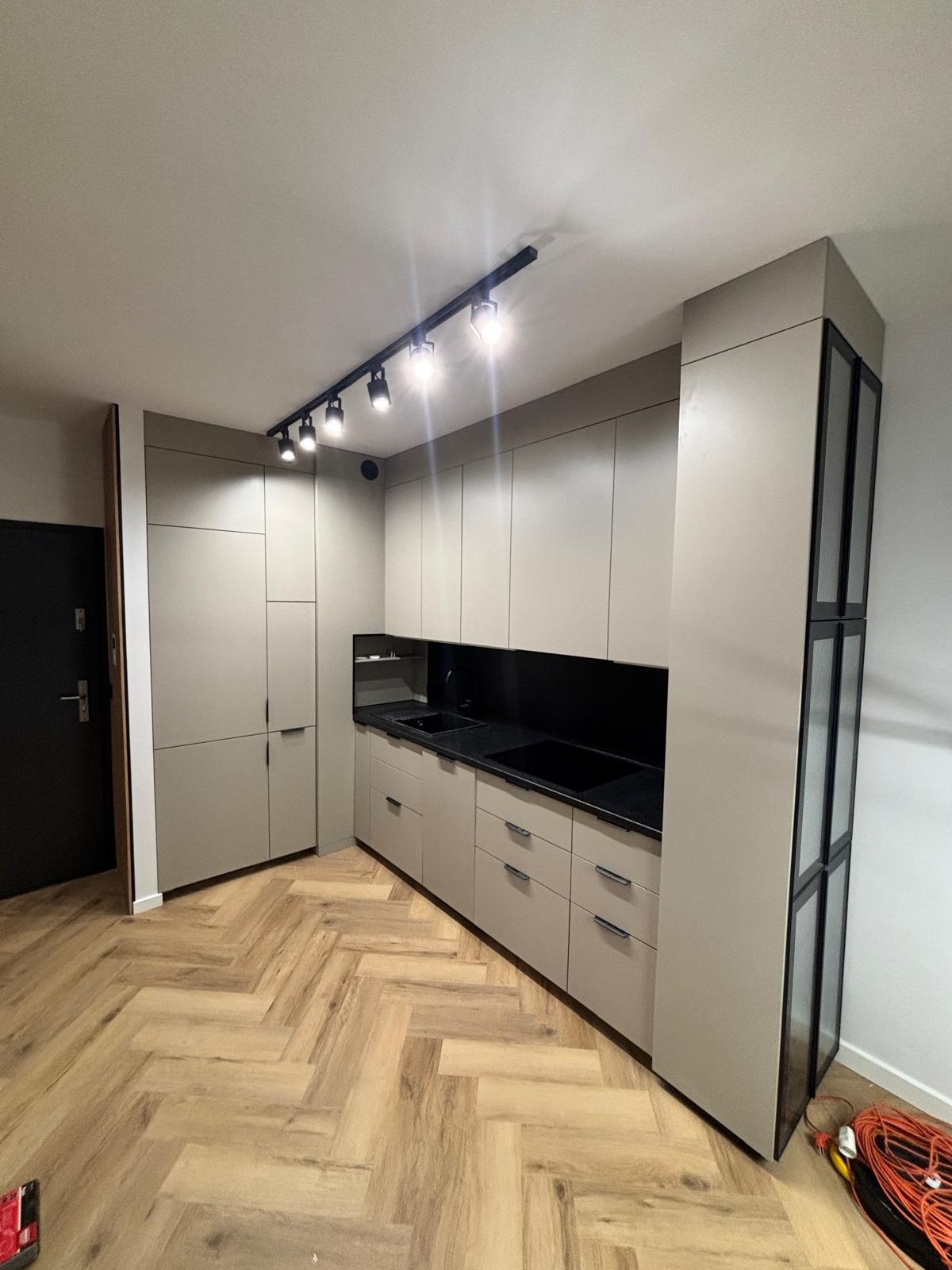 Nowoczesna kuchnia w odcieniach beżu z czarnymi akcentami, oświetleniem punktowym i parkietem ułożonym w jodełkę. Widoczna zabudowa kuchenna i drzwi wejściowe.