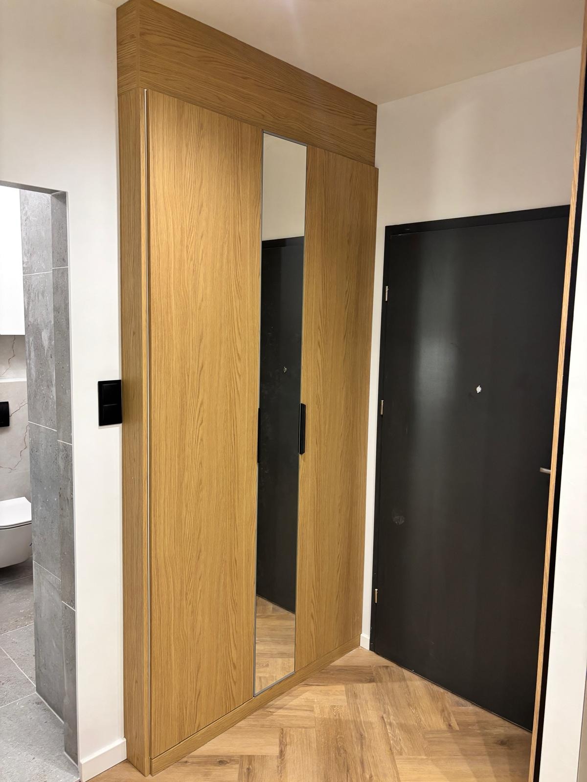Drewniana zabudowa szafy z lustrem w przedpokoju, widok na podłogę w jodełkę i czarne drzwi. Minimalistyczny design, jasne wnętrze.