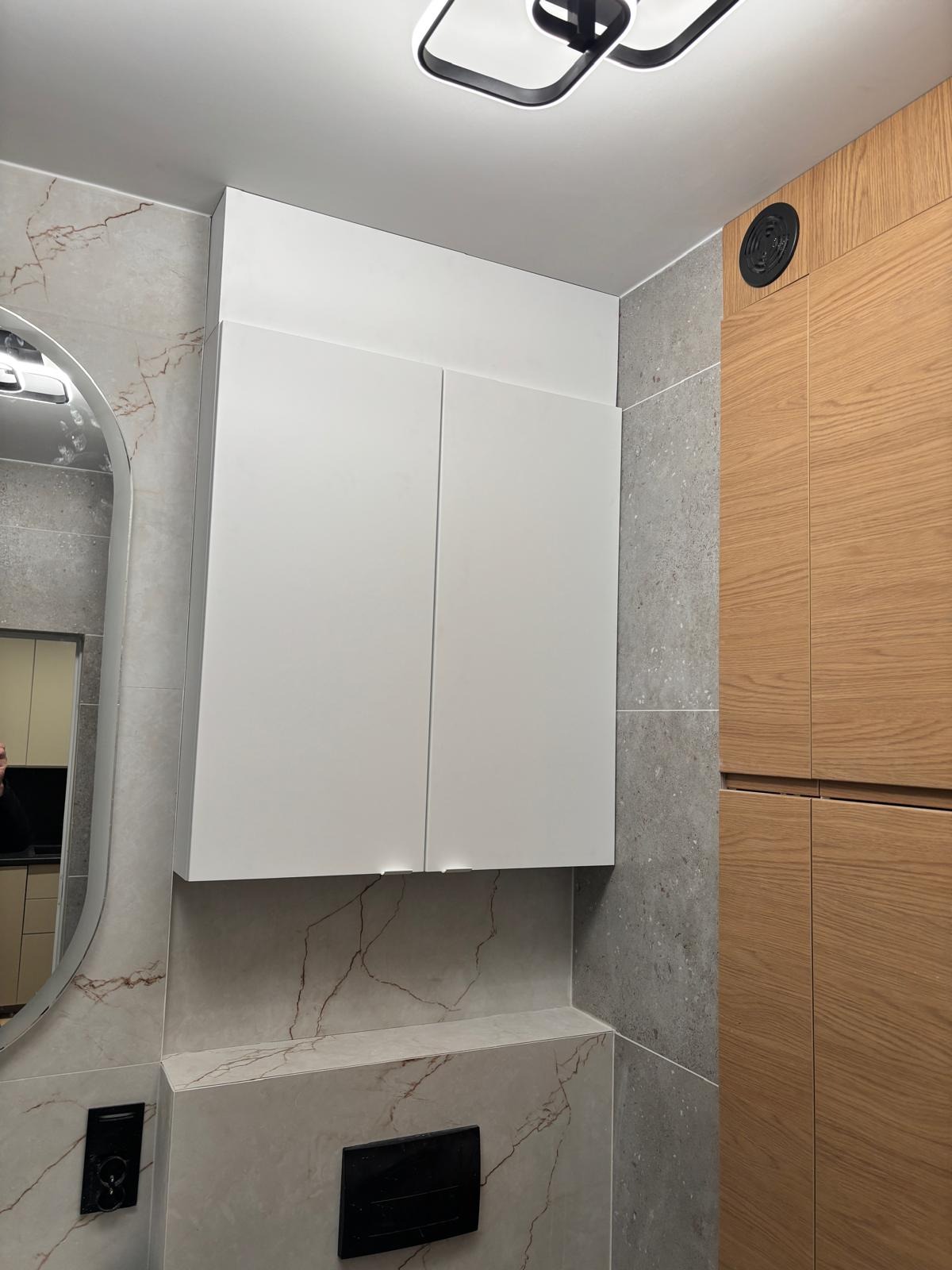 Nowoczesna łazienka z szafką nad toaletą, płytkami imitującymi marmur i drewnianą zabudową. Minimalistyczny design, oświetlenie sufitowe.