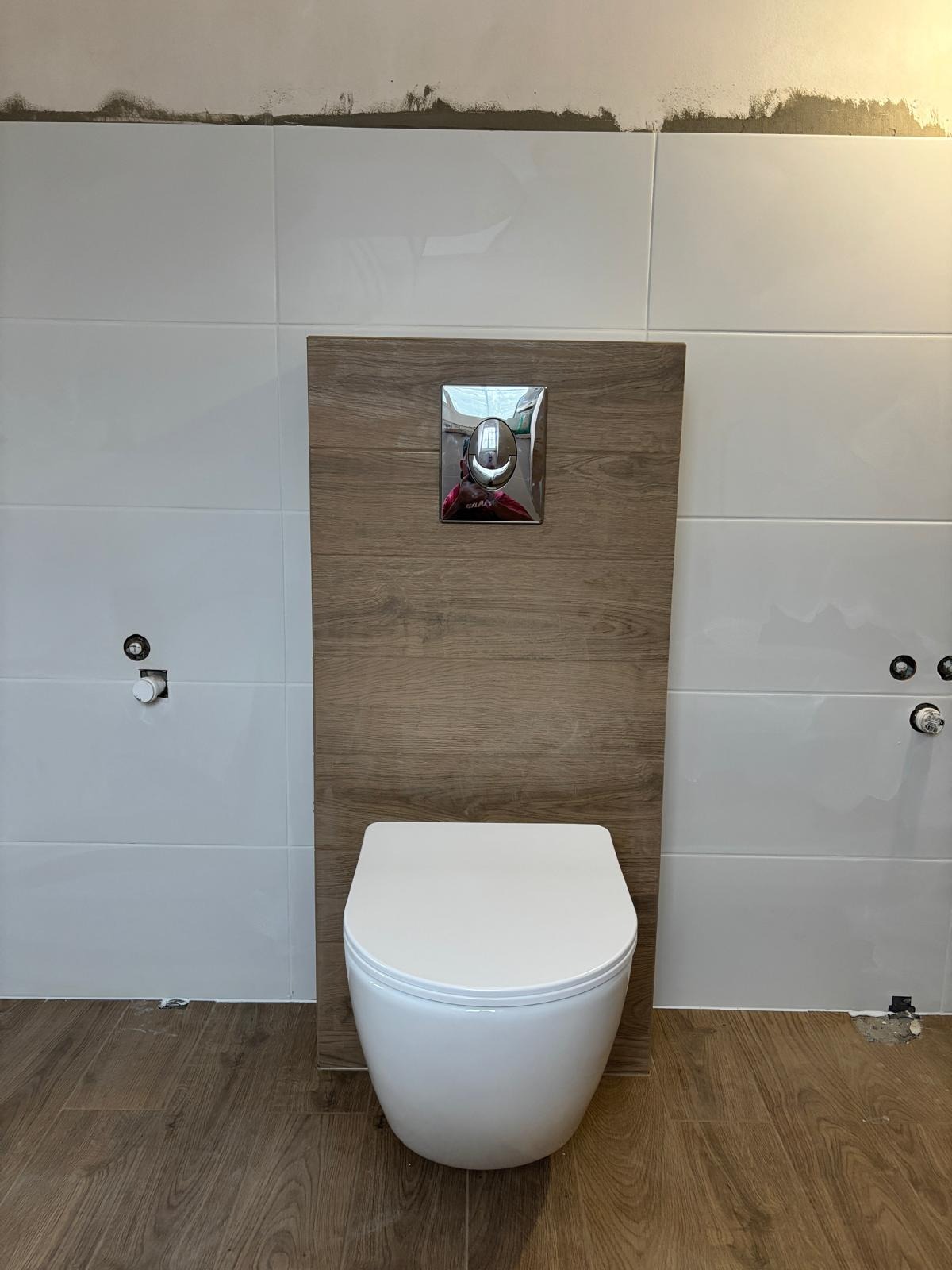 Nowoczesna toaleta podwieszana z drewnianą obudową i chromowanym przyciskiem spłukującym, białe płytki na ścianach i podłoga imitująca drewno. Wykończenie łazienki.